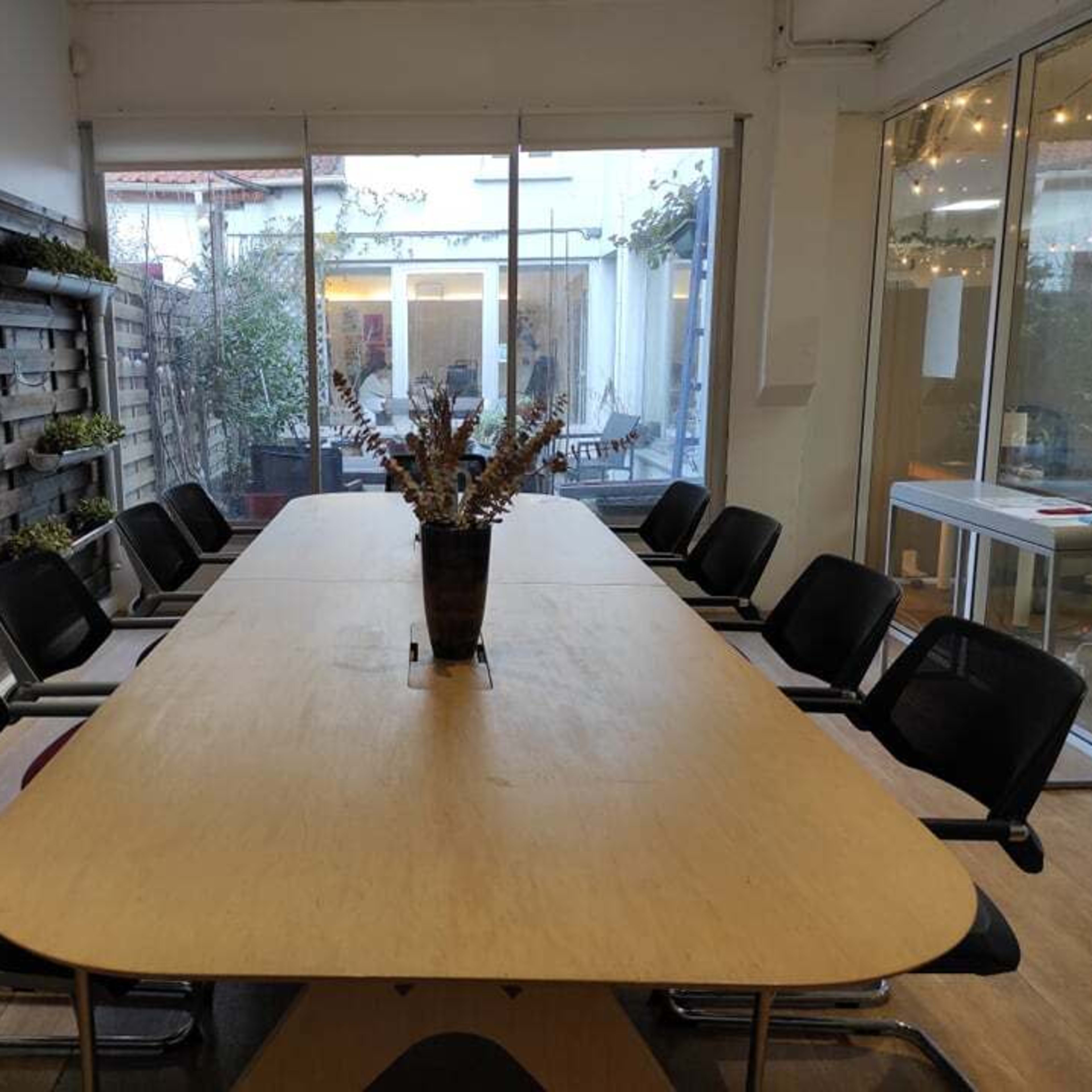 A large wooden conference table is set in a well-lit room with chairs around it, overlooking an outdoor patio through glass walls.