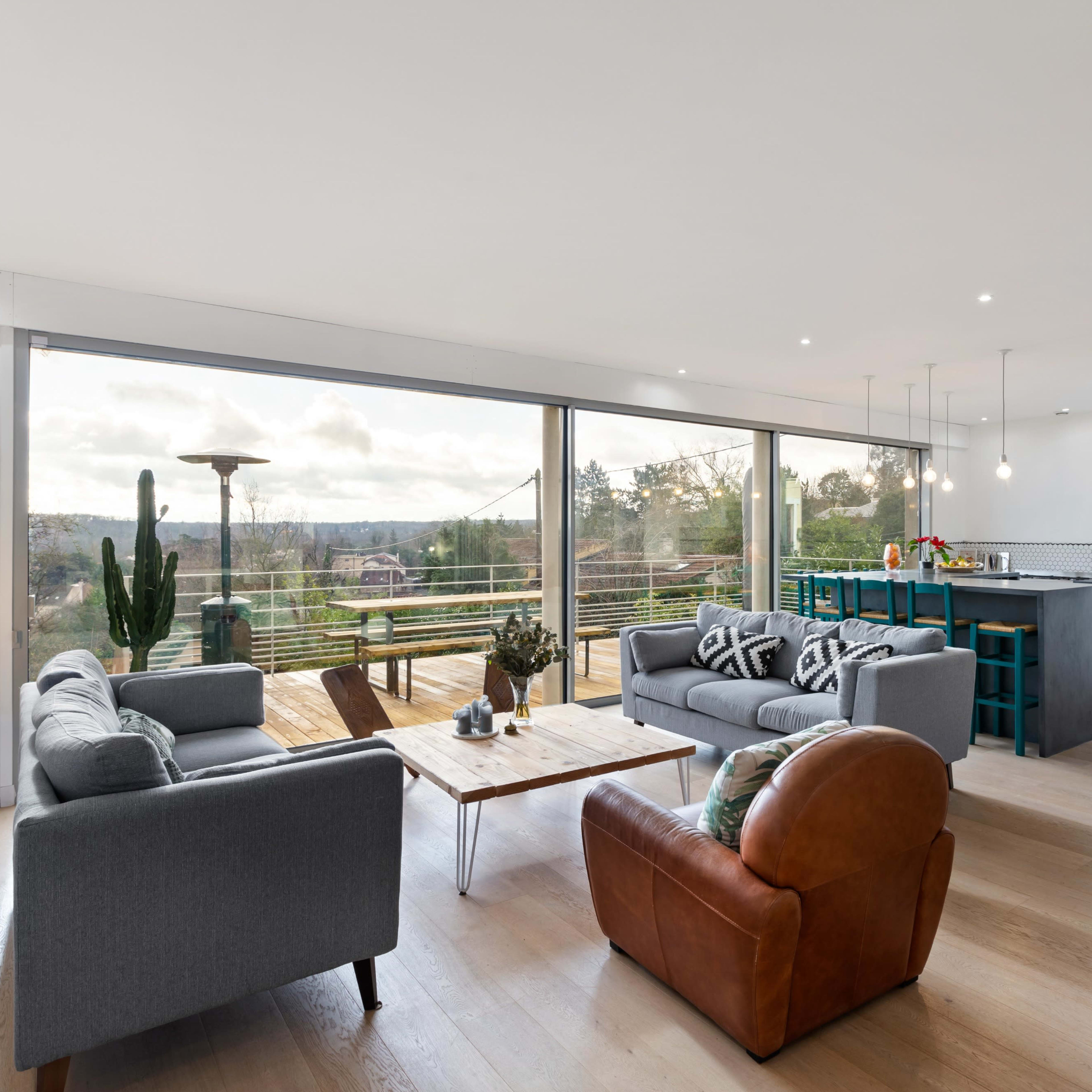 A modern living room with two gray sofas, an armchair, a coffee table, and a kitchen visible in the background, featuring large windows that overlook a scenic view.