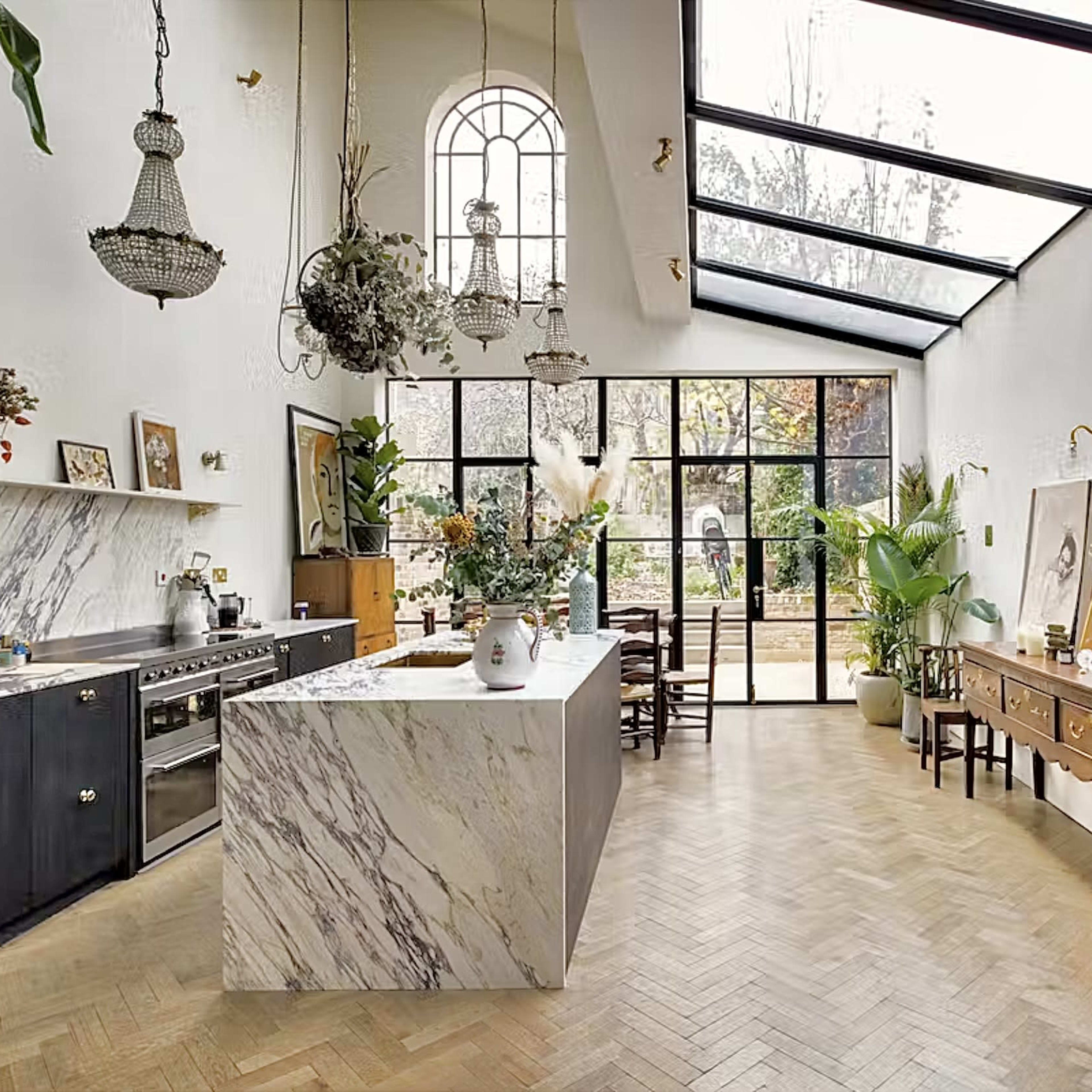 A spacious kitchen and dining area with a large marble island, high ceilings, and large glass doors leading to a garden.