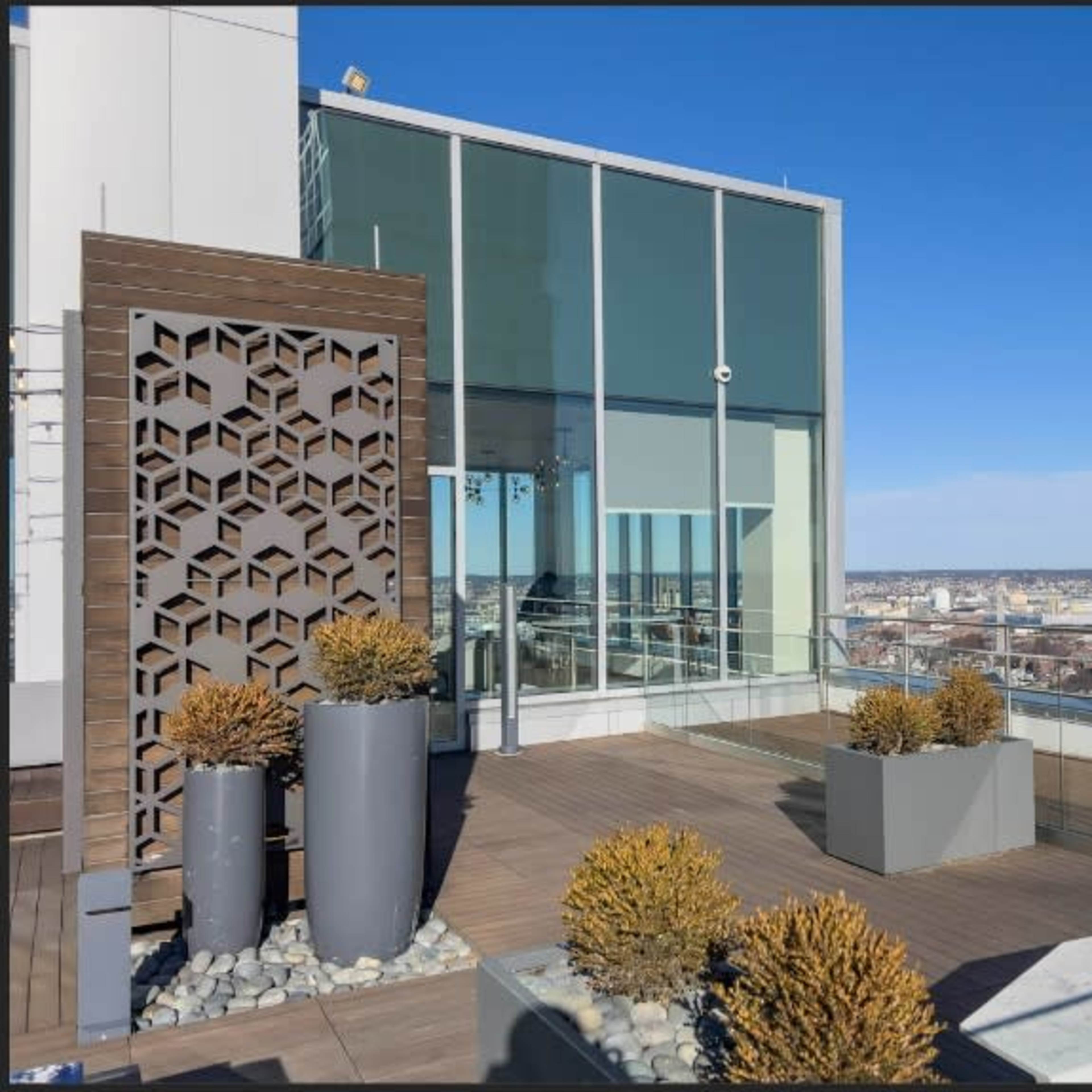 A modern rooftop terrace featuring decorative planters, a geometric wooden screen, and a panoramic view of a city skyline under clear blue skies.