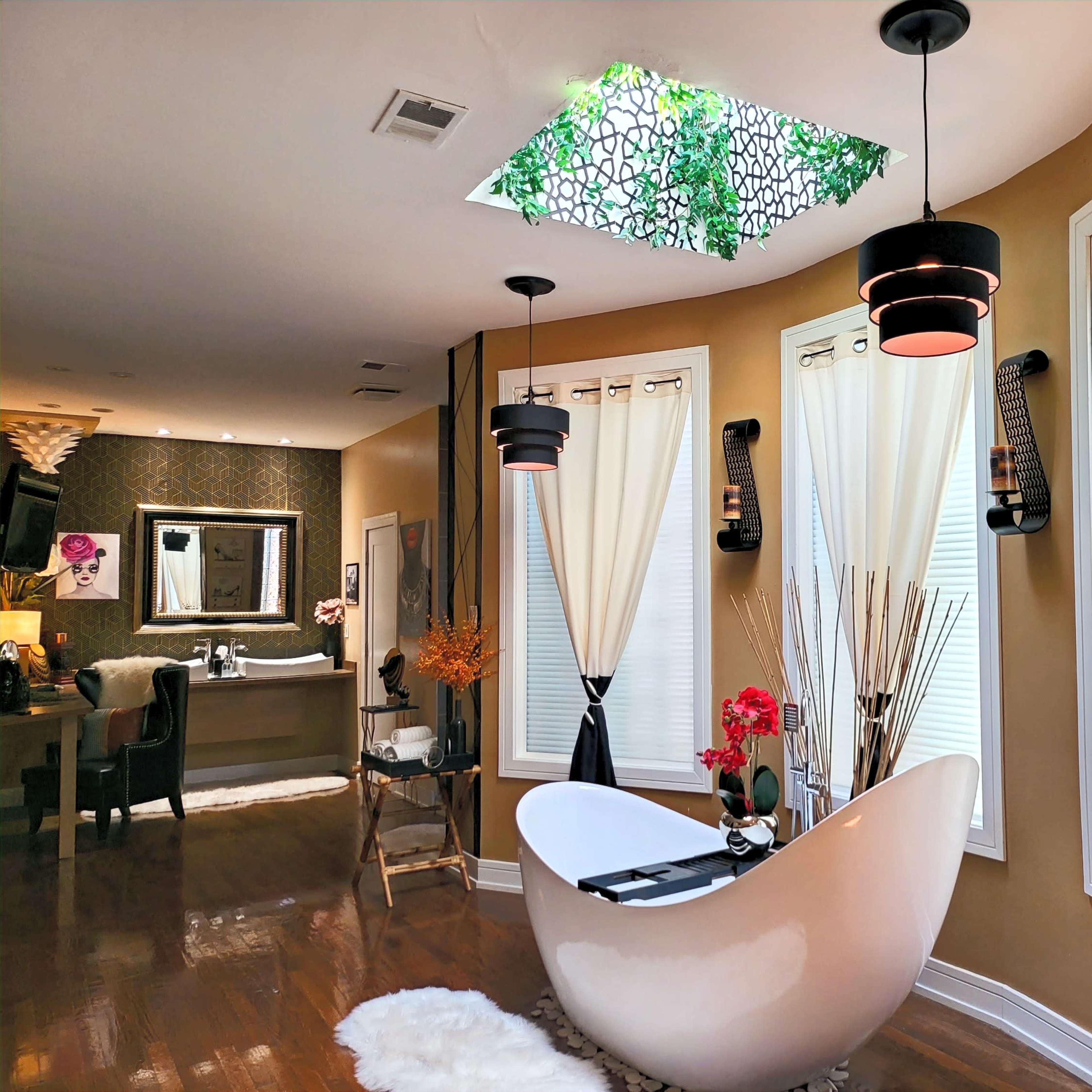 A modern bathroom featuring a freestanding bathtub surrounded by large windows, with light curtains and decorative plants in the ceiling skylight.
