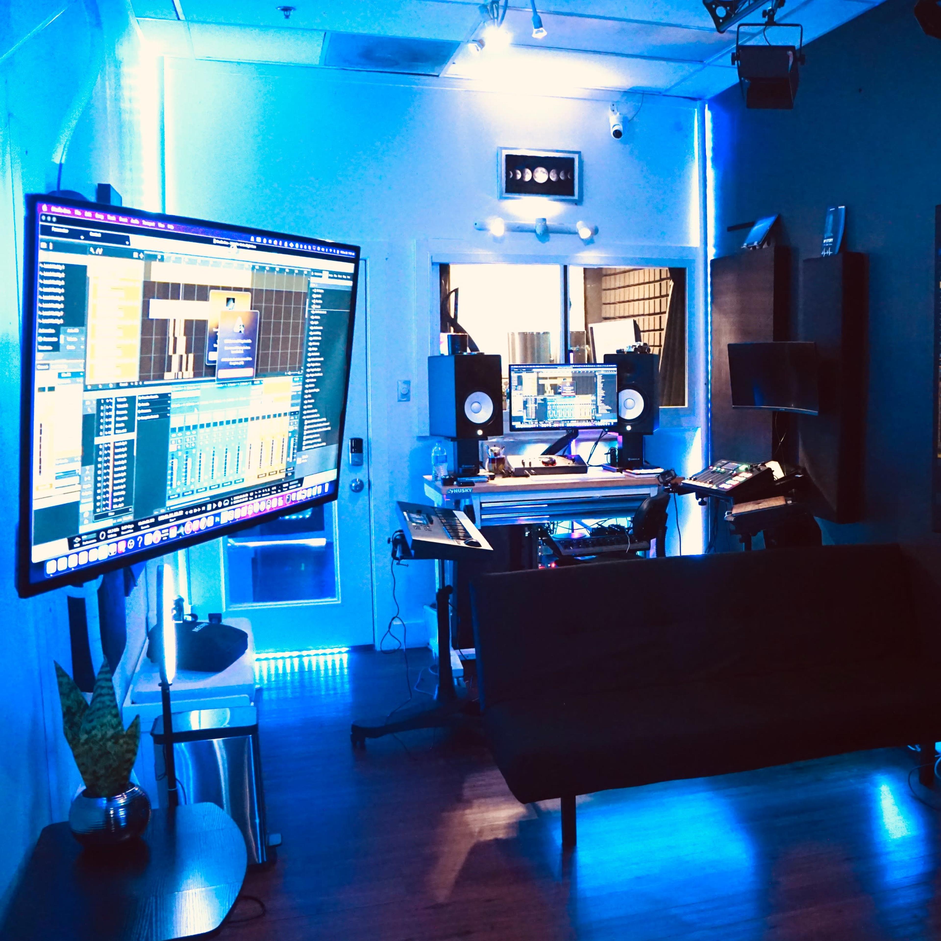 A recording studio with multiple large screens displaying audio software, a production desk, and a seating area.
