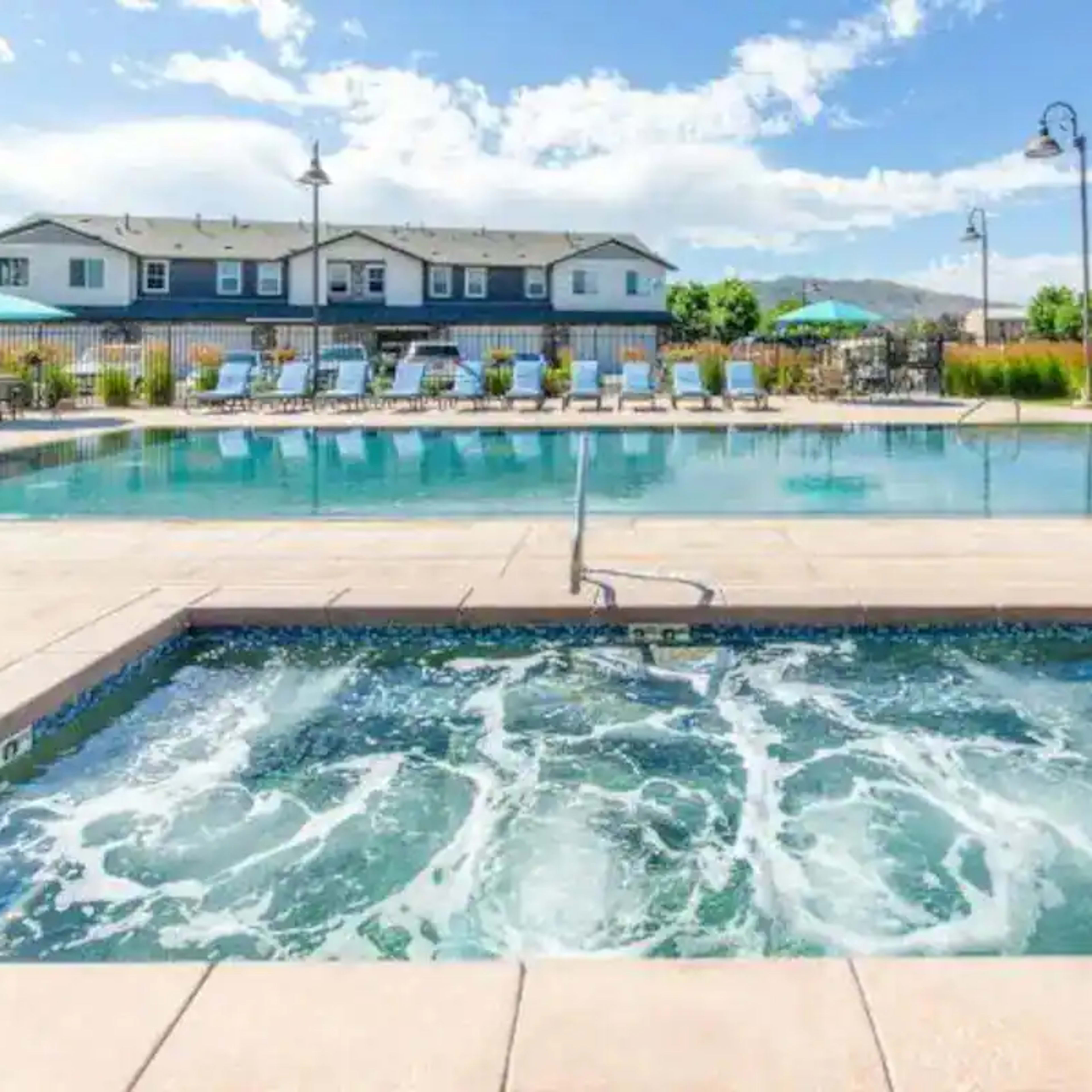 A residential pool area featuring a large swimming pool and a hot tub, surrounded by lounge chairs and umbrellas.