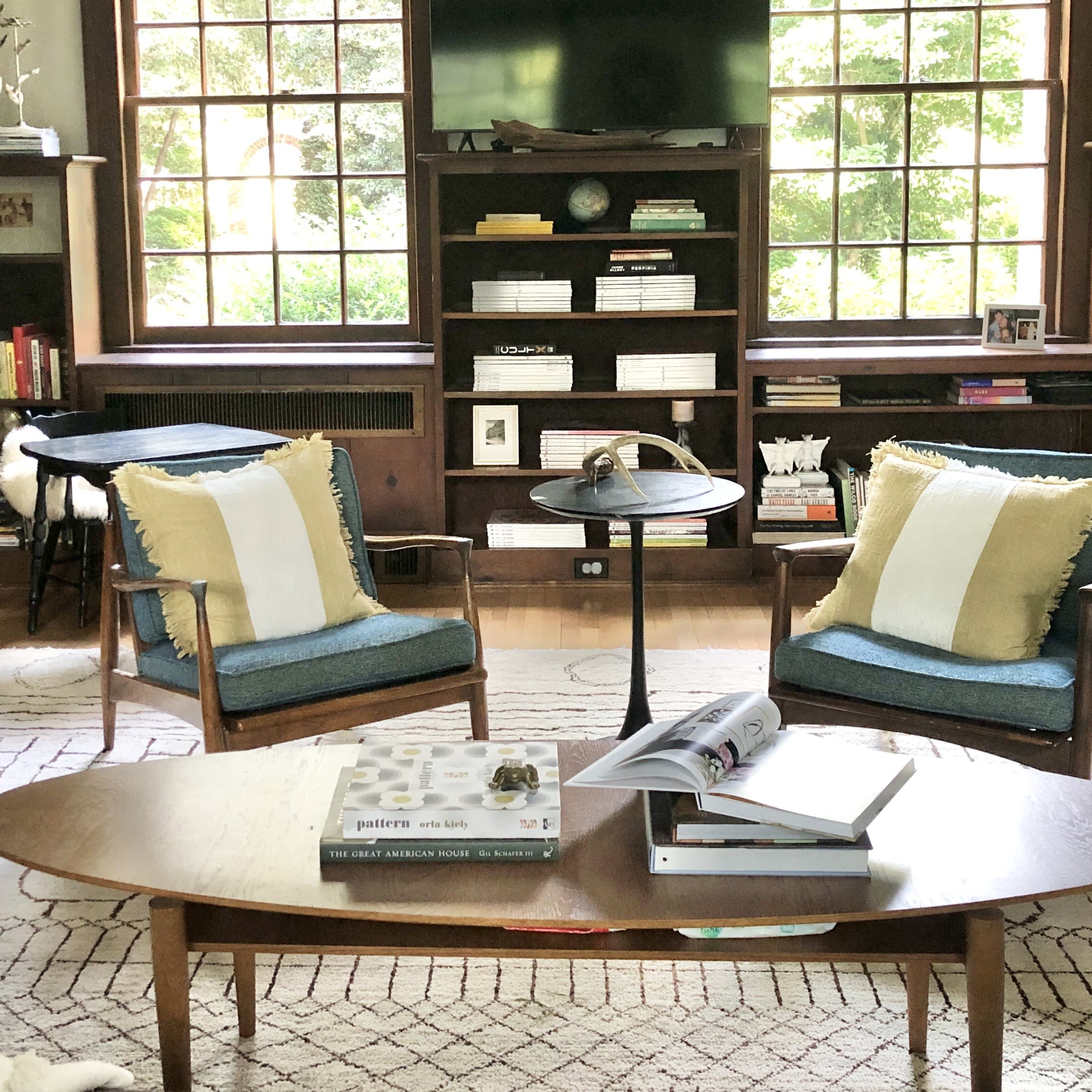 A living room features two chairs with cushions, a coffee table with books, and a large window overlooking greenery.