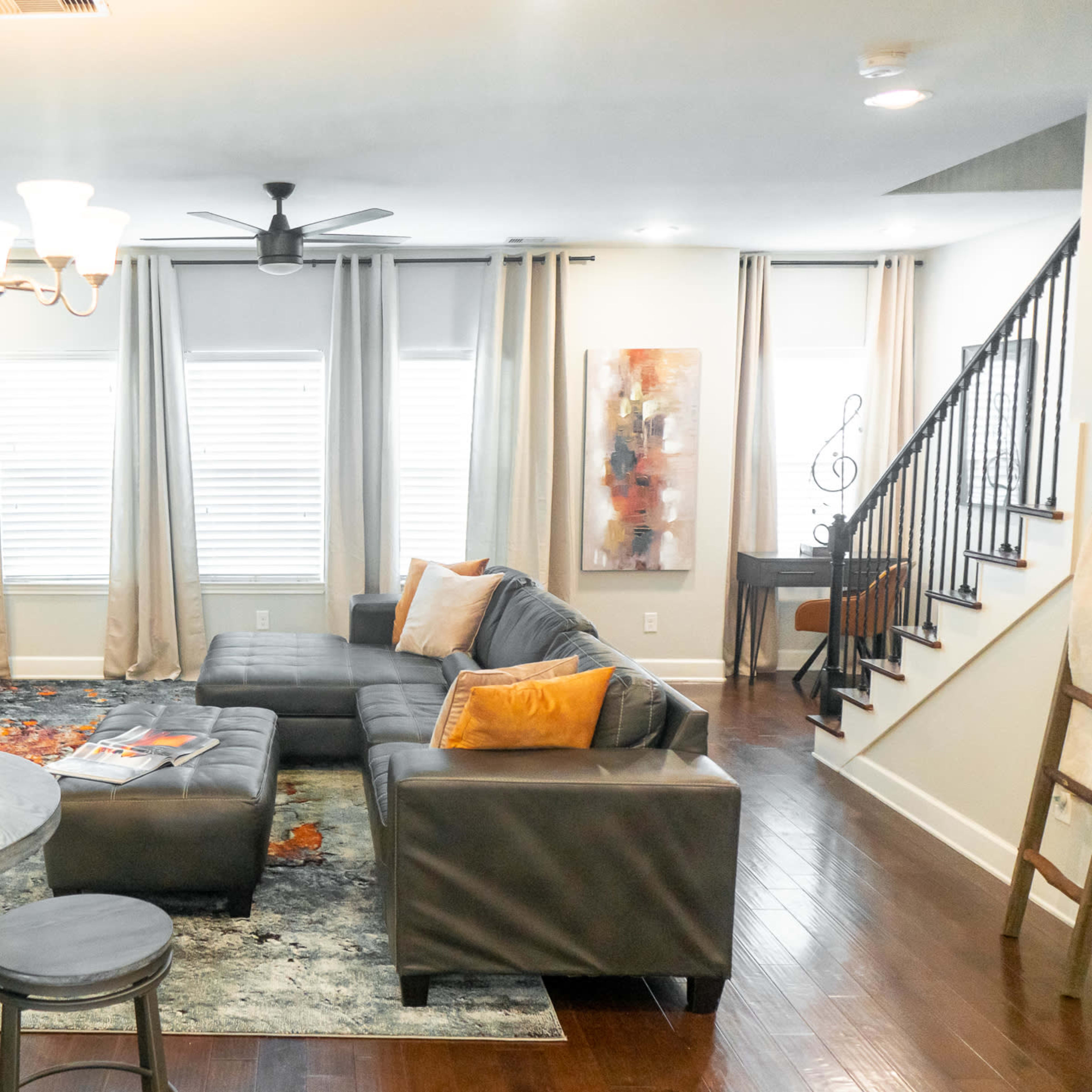 A modern living room features a sectional sofa, a dining table, large windows with curtains, and a staircase in the background.