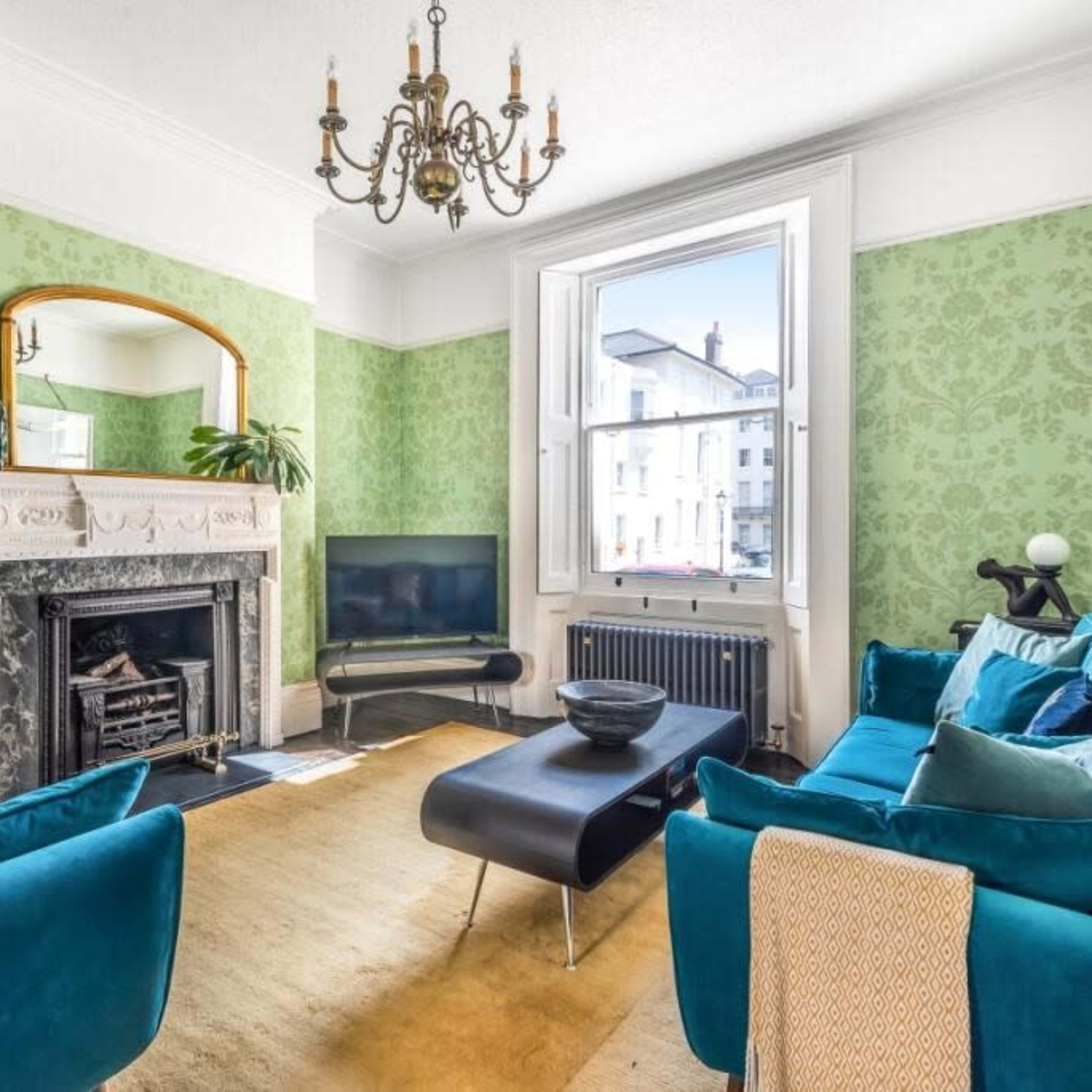 The living room features green wallpaper, a fireplace with a marble surround, a large window, and turquoise sofas arranged around a black coffee table.