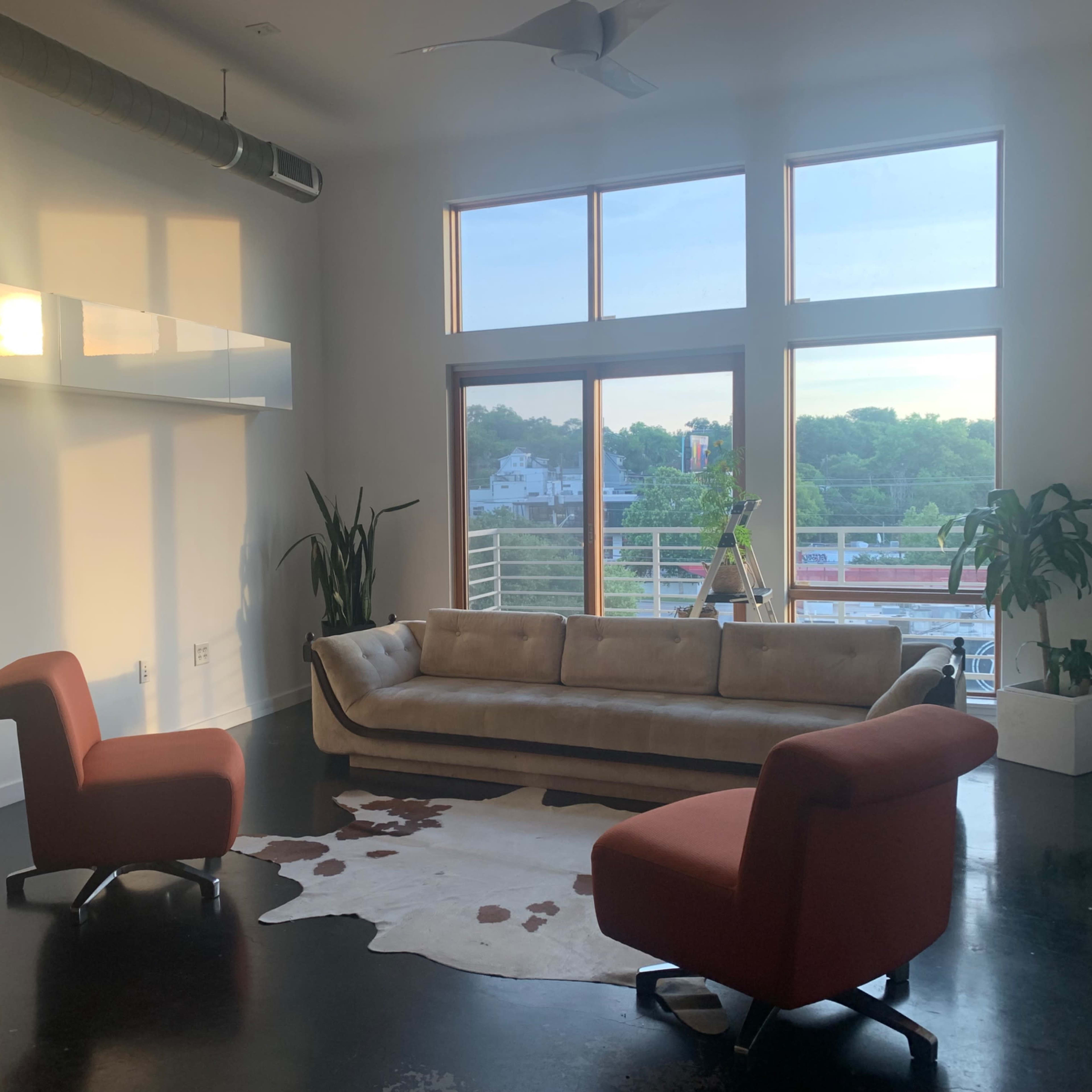 A spacious living room features a beige sofa, two red chairs, and large windows that overlook a cityscape.