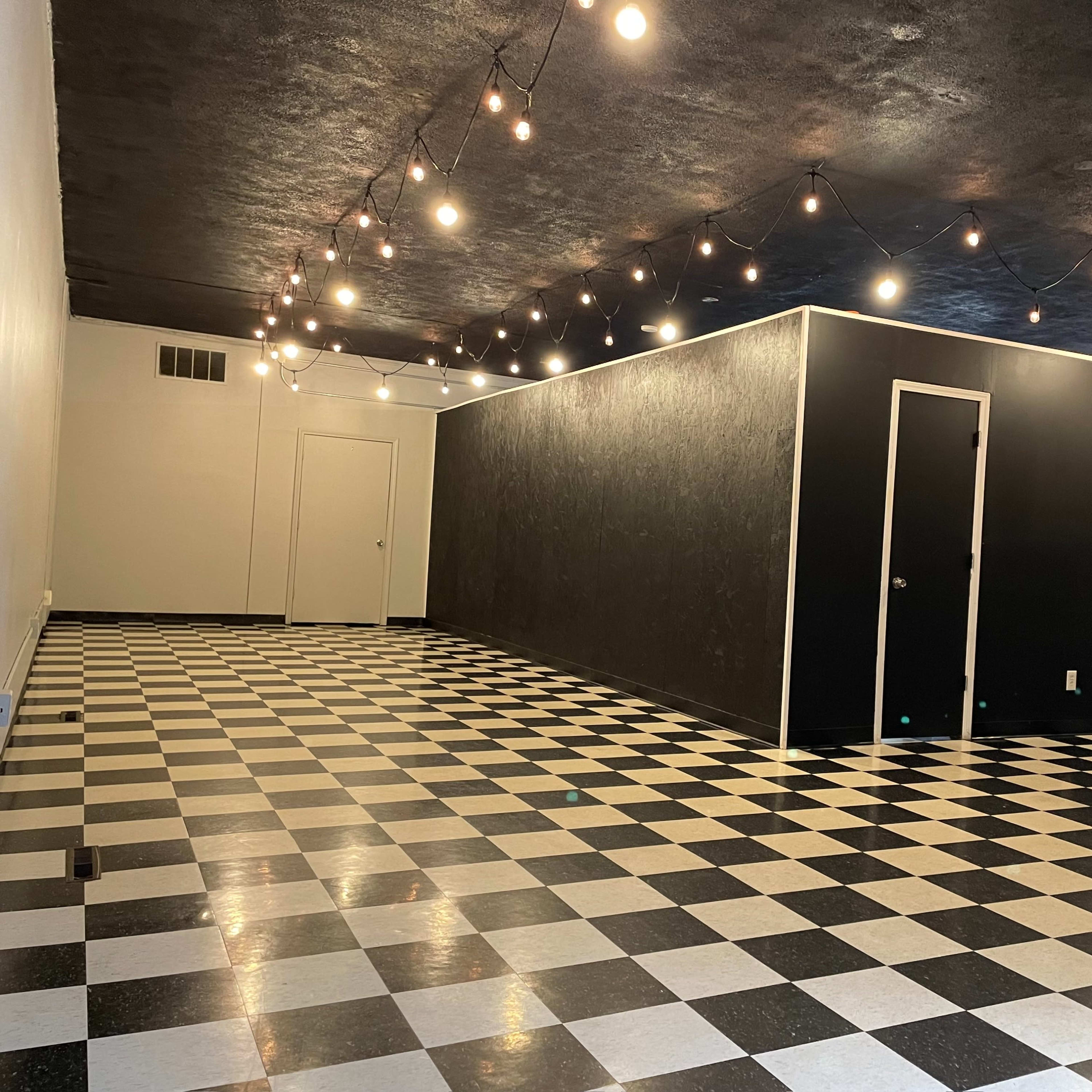 The room features a black and white checkerboard floor, string lights hanging from the ceiling, and a black accent wall with a door.
