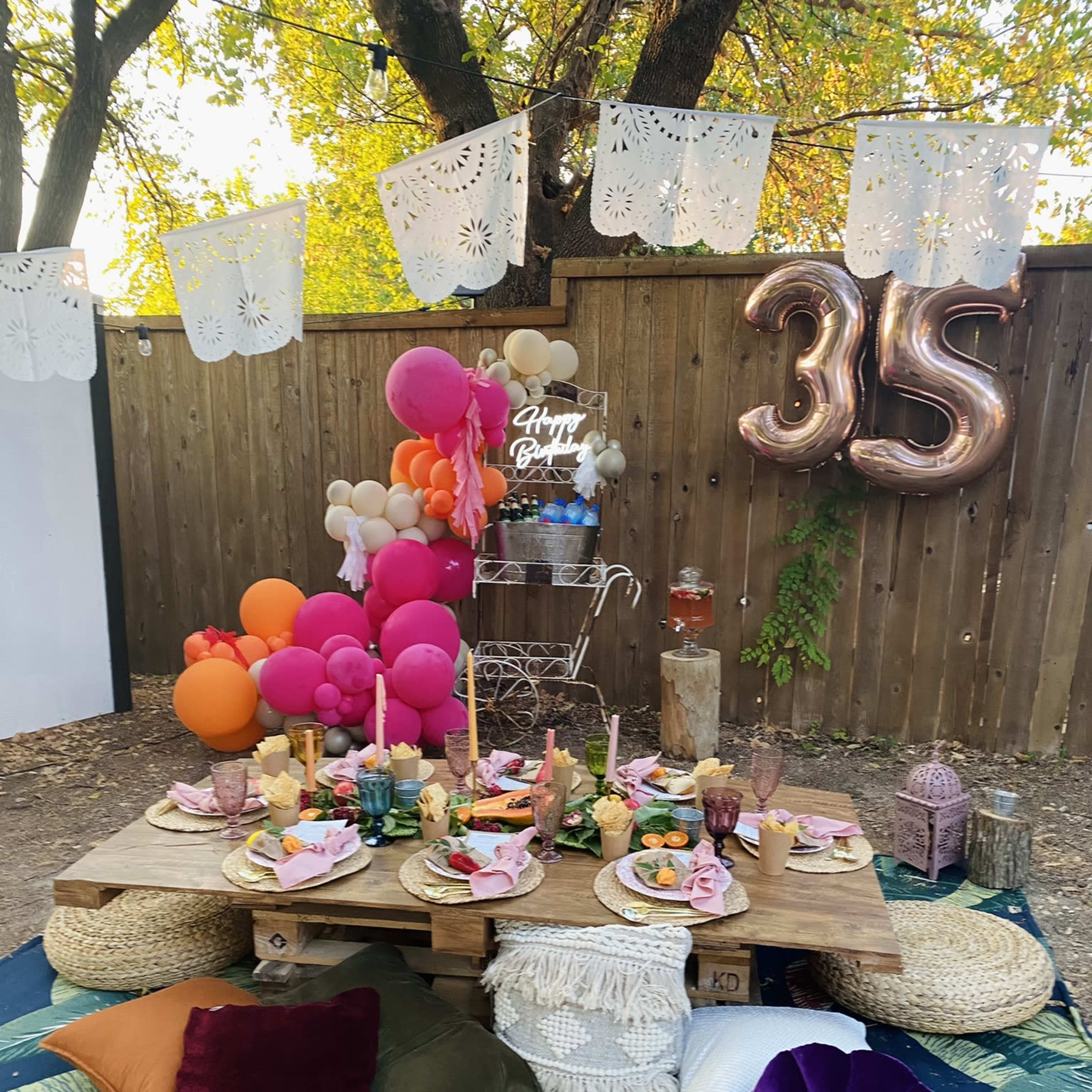 A backyard party setup features a low wooden table adorned with colorful dishes and drinks, surrounded by decorative balloons and a large "35" balloon, set on a layered rug with pillows.