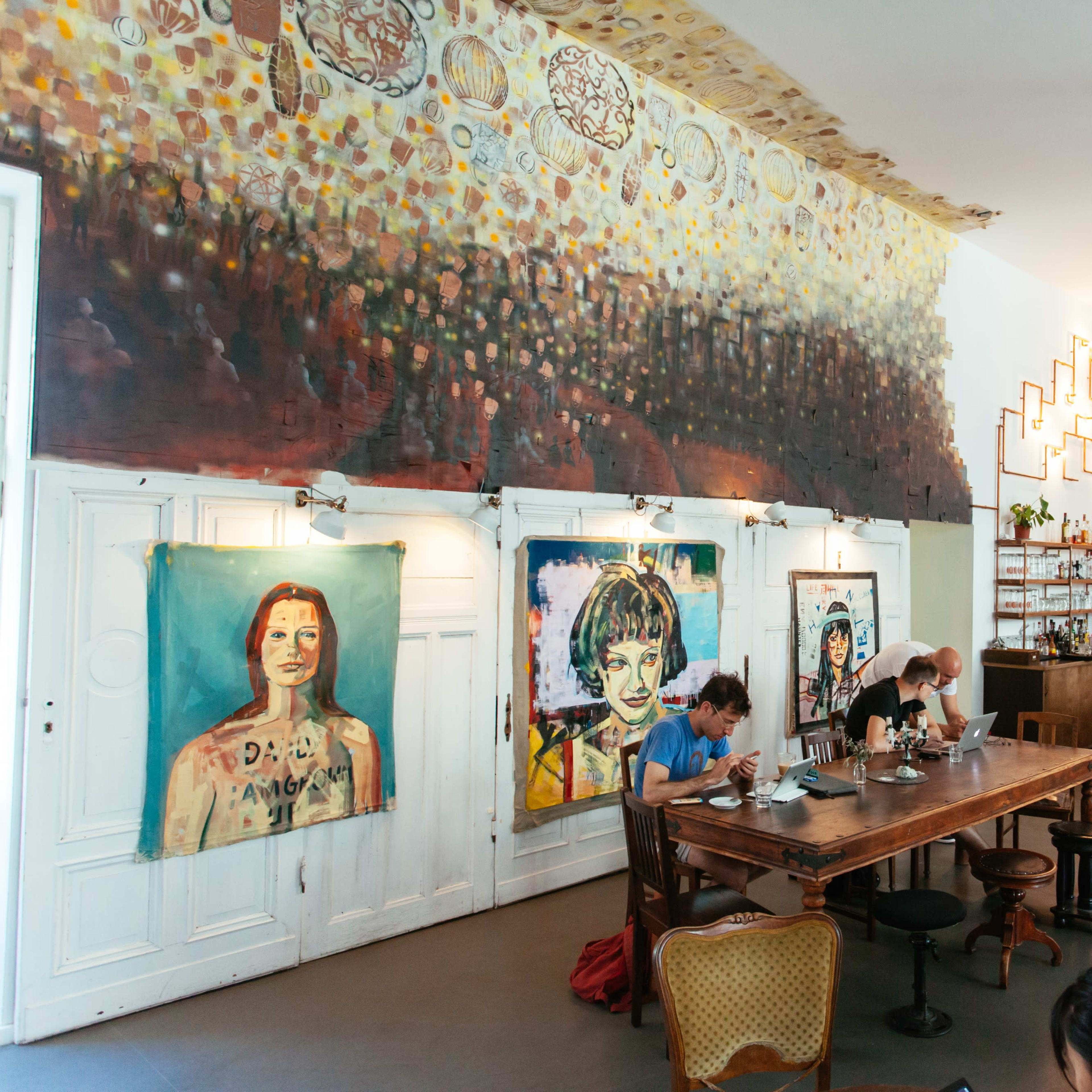 A café interior featuring large colorful paintings on the walls, a wooden dining table, and customers sitting at tables and the bar.