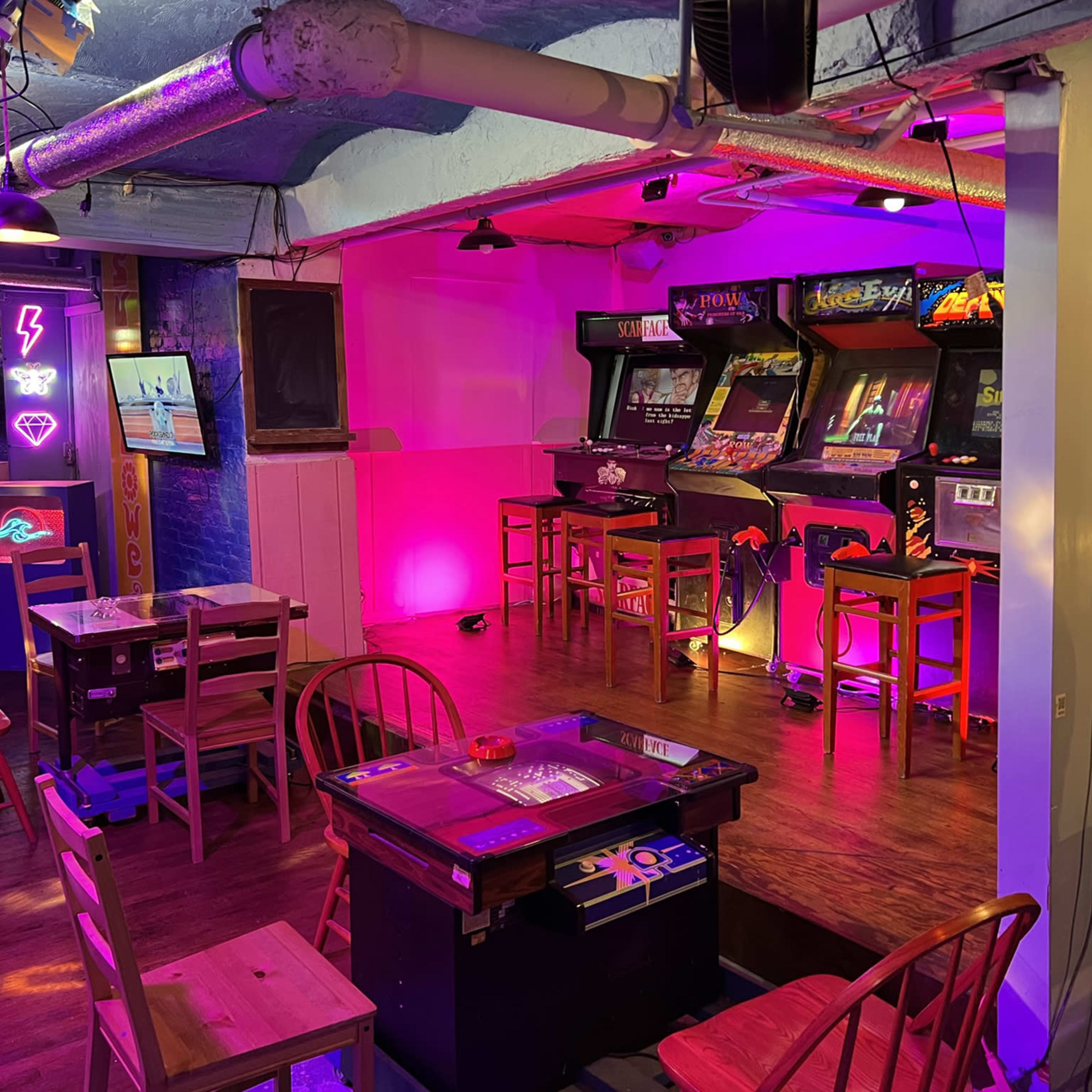 A retro arcade with multiple video game machines and tables, illuminated by colorful lights.