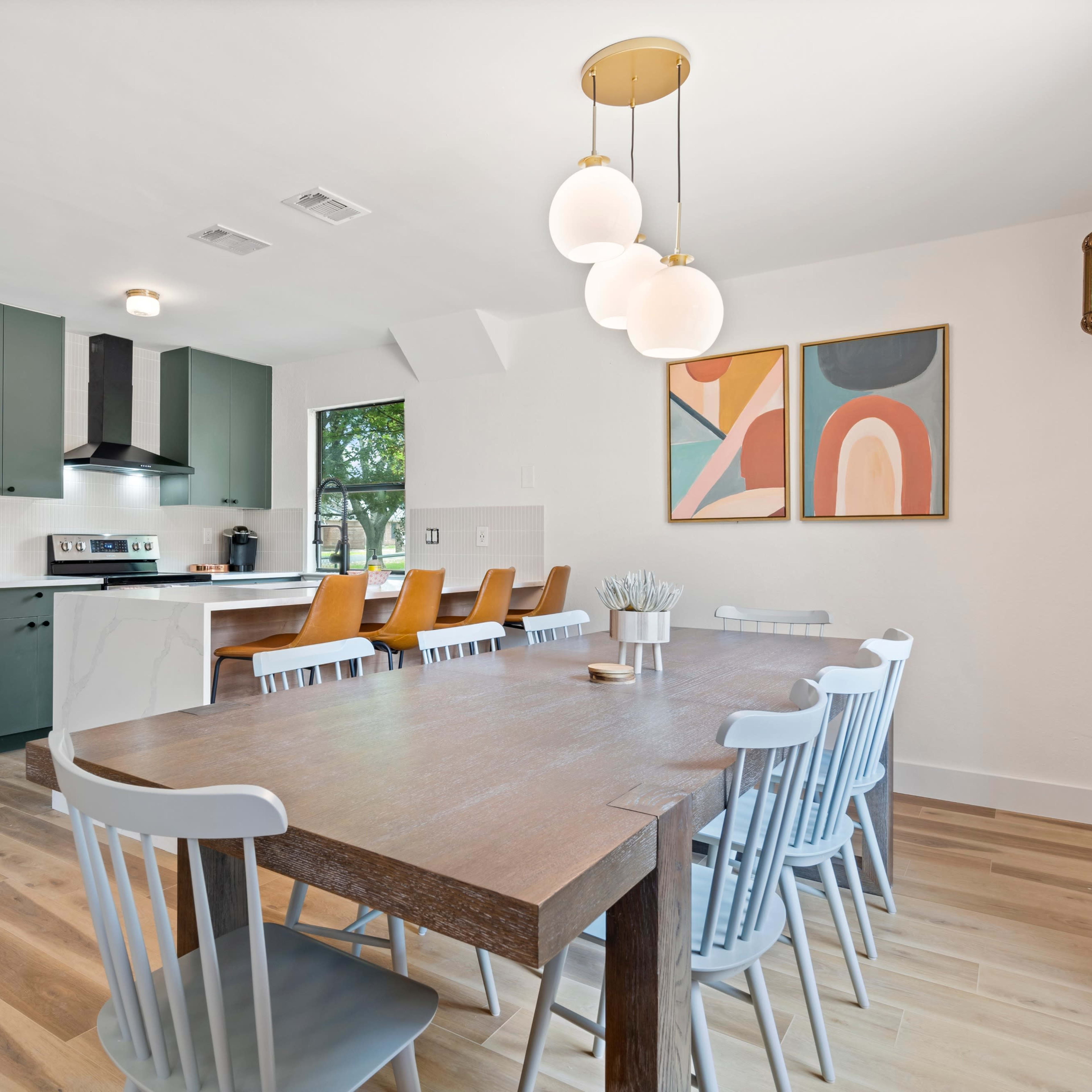 A large dining table with light gray chairs is positioned in a modern kitchen featuring dark green cabinets, a white countertop, and art on the walls.