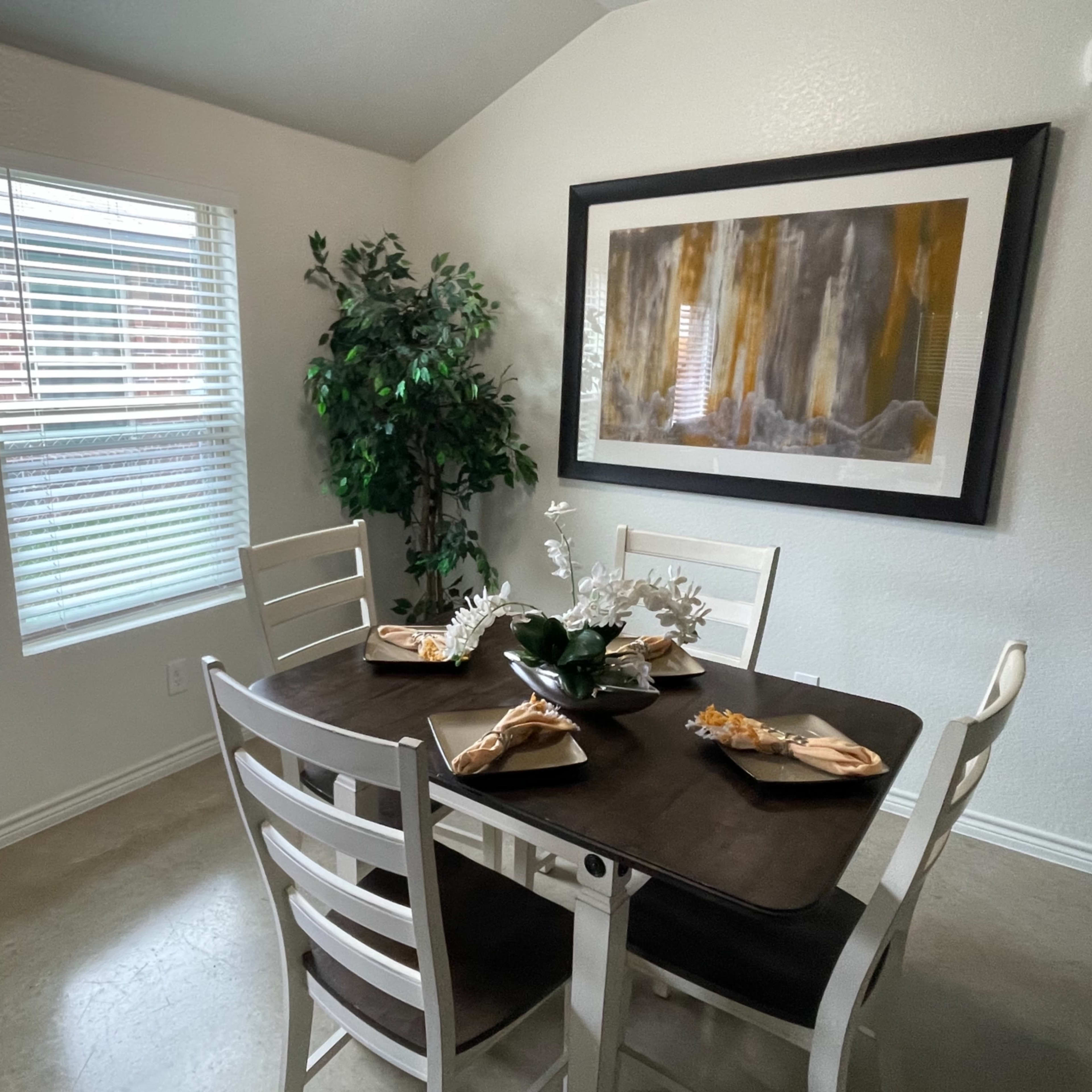 A dining area with a brown table set for four, adorned with decorative plates and a centerpiece, alongside a window and a potted plant.