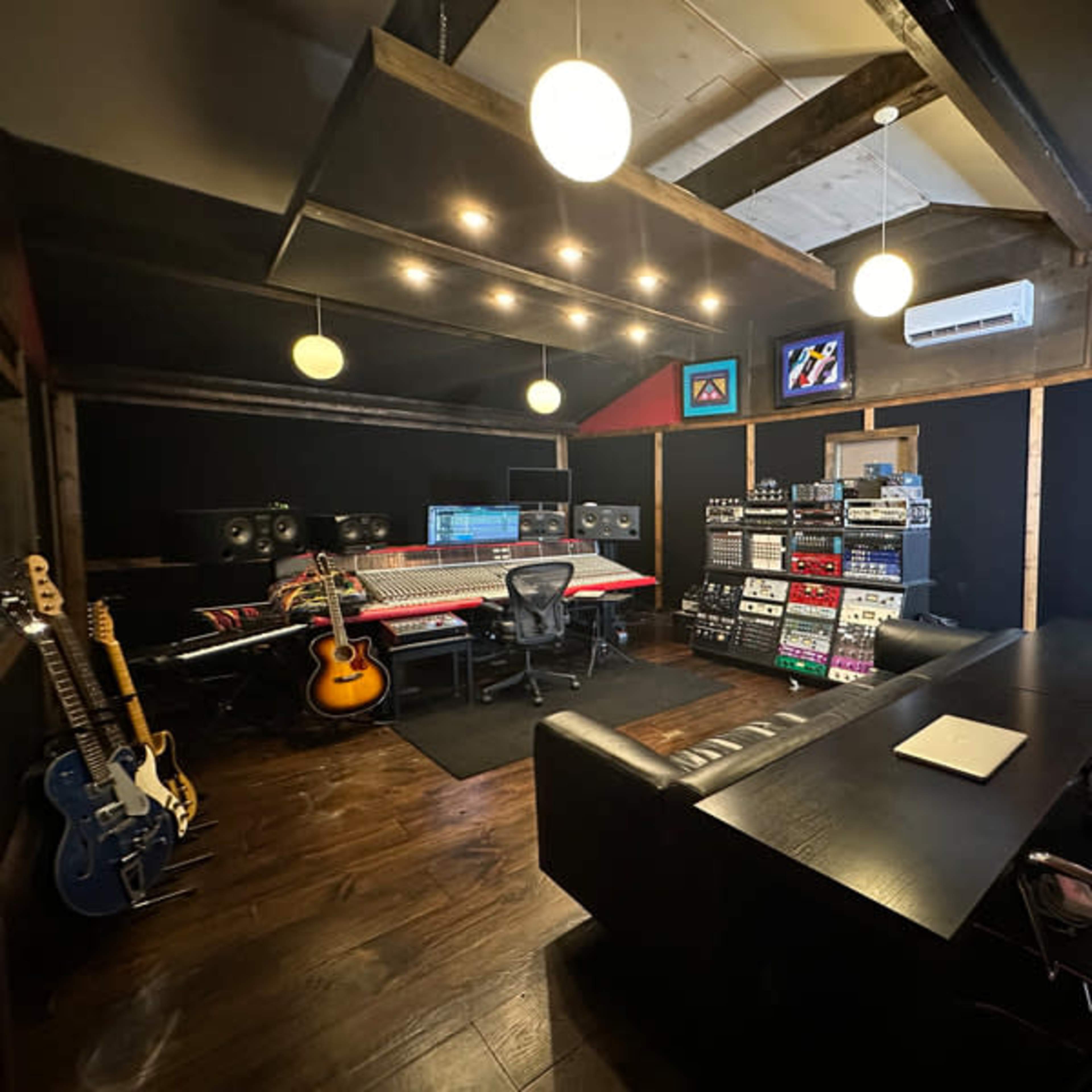 A music production studio featuring a central mixing console, audio equipment racks, several guitars, and a conference table with chairs.