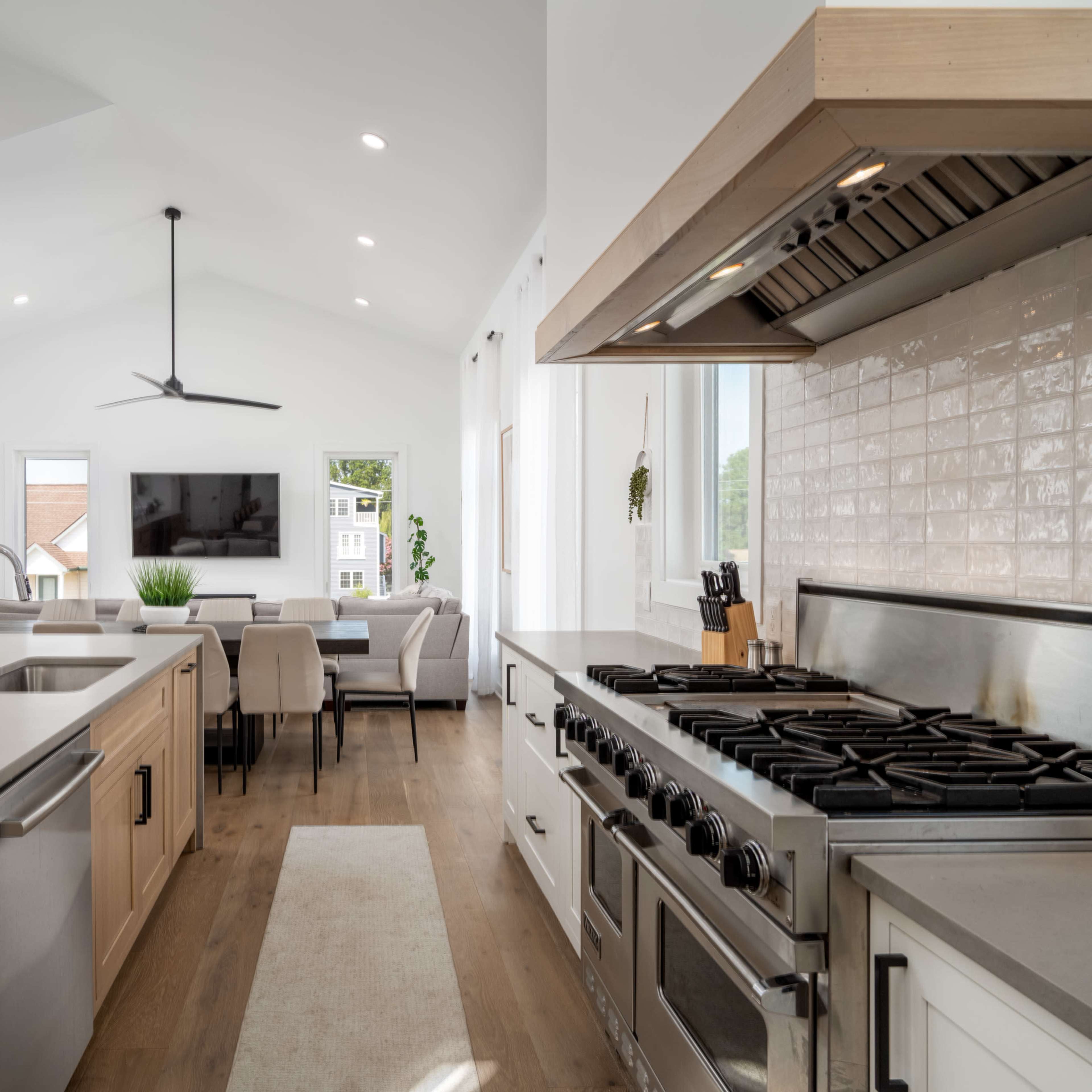 A modern kitchen with wooden cabinets, a long countertop, and a professional-grade stove opens up to a dining area and living space with large windows.