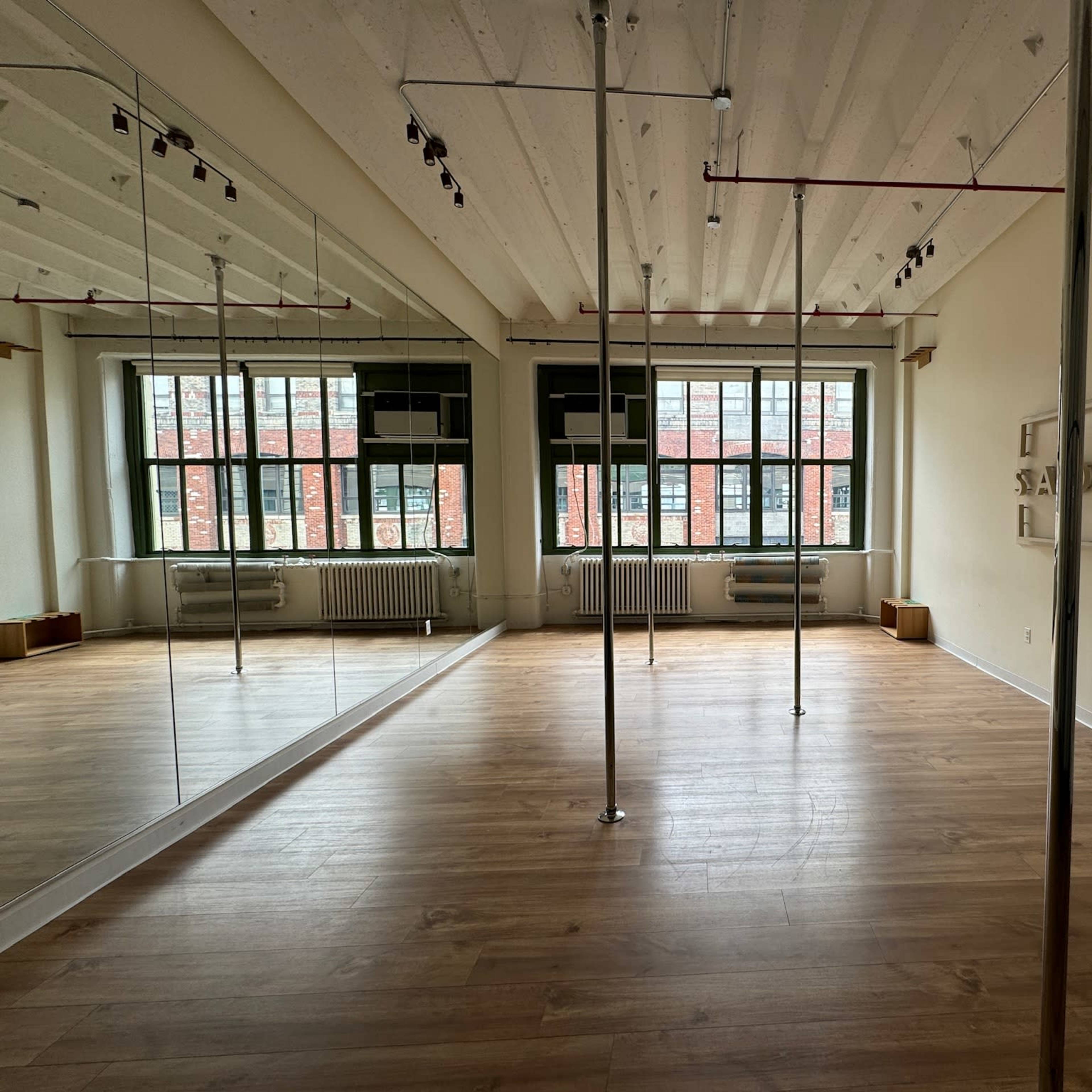 A spacious, empty room with polished wooden floors, large windows, and mirrored walls, featuring two vertical poles in the center.