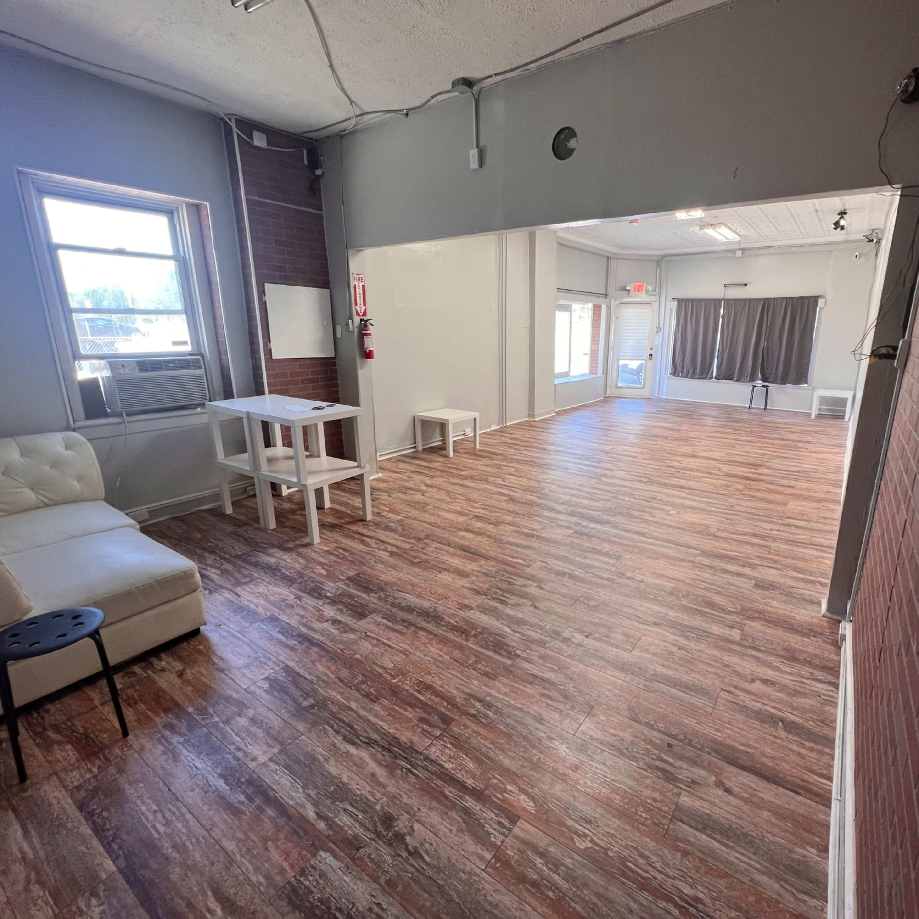 A spacious room with a wooden floor, a white couch, a small table, and large windows that allow natural light to enter.