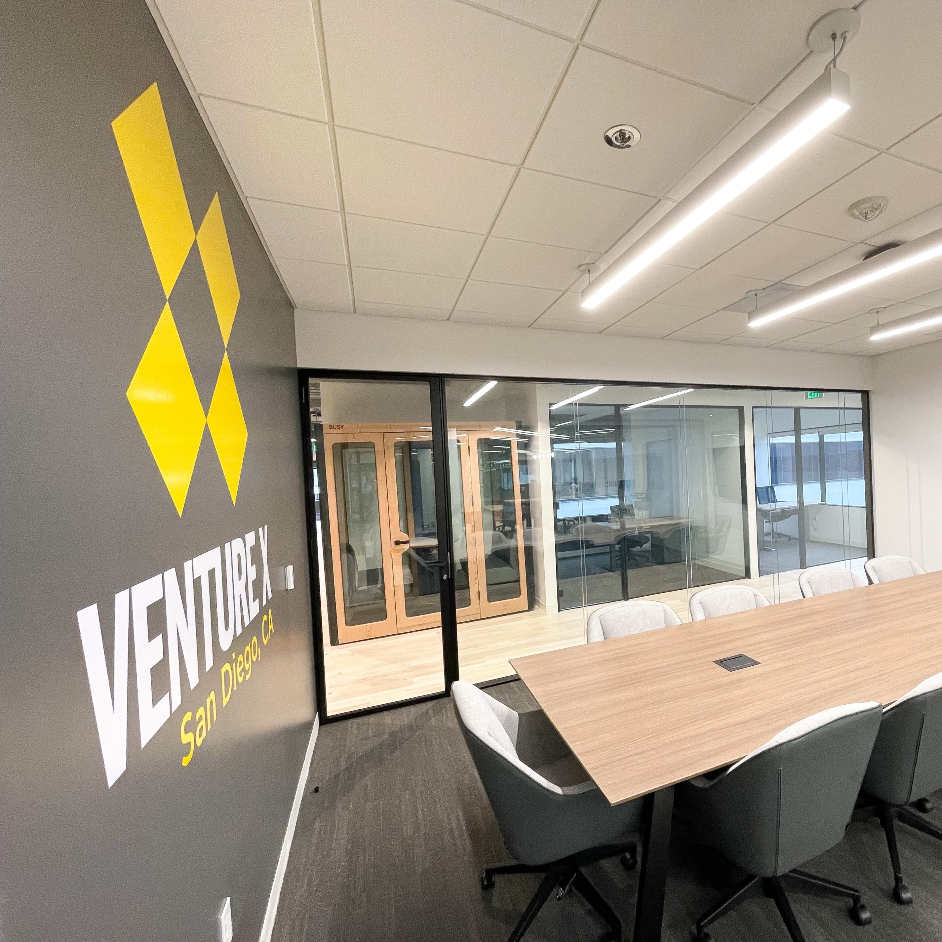 A spacious conference room features a long wooden table, ergonomic chairs, and glass walls with a logo displayed on the adjacent wall.