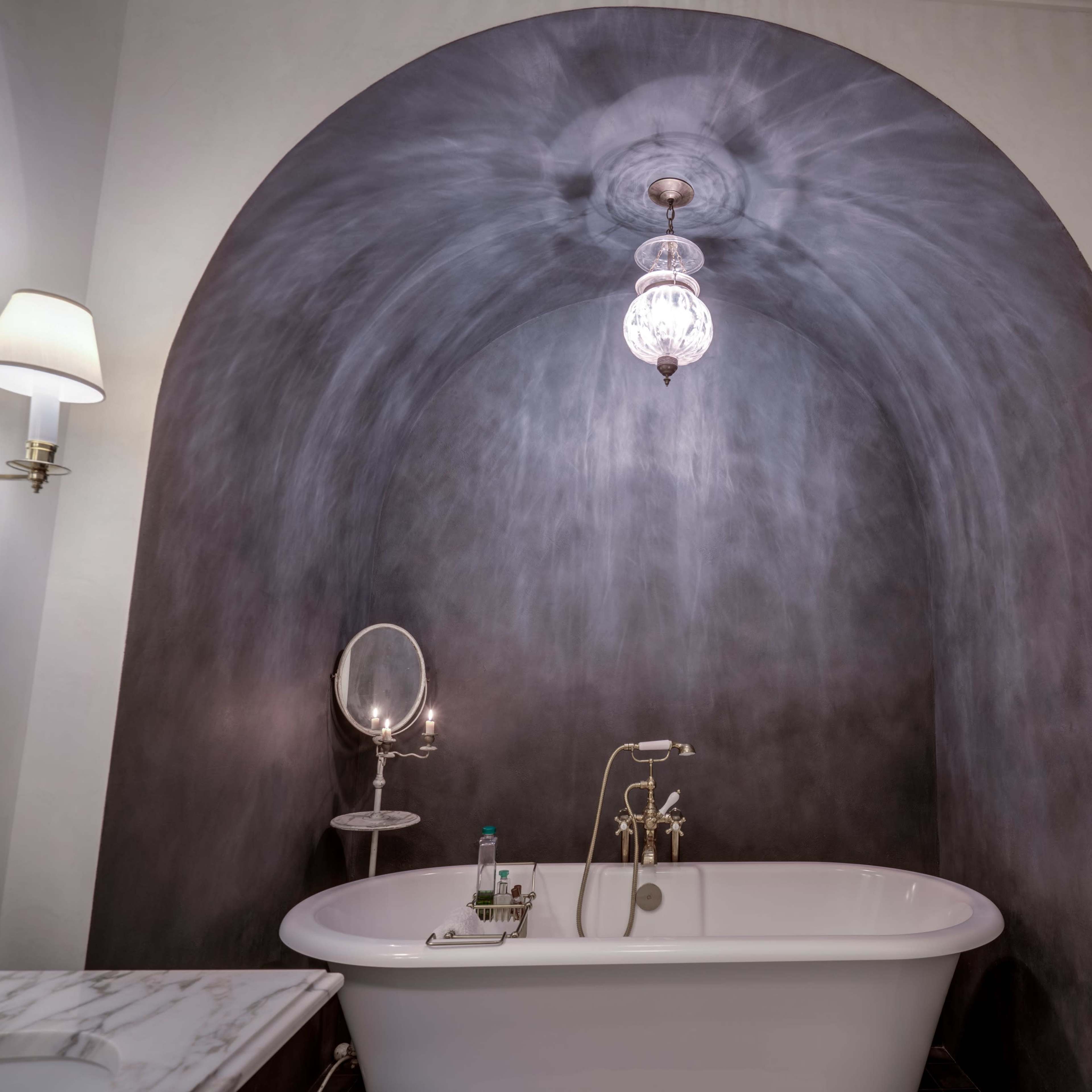 The scene features a freestanding bathtub positioned against a curved gray wall, illuminated by a decorative light fixture.