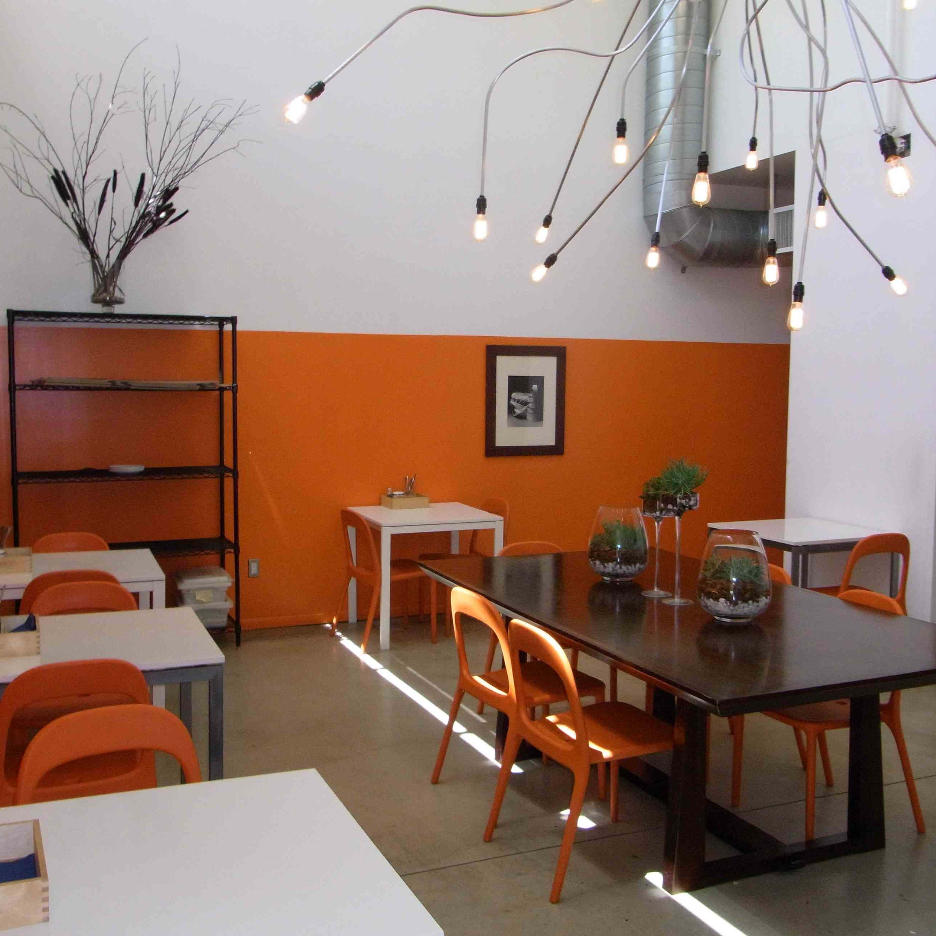 A modern workspace featuring orange accent walls, a large wooden table, and several white tables and chairs, all under unique pendant lighting.