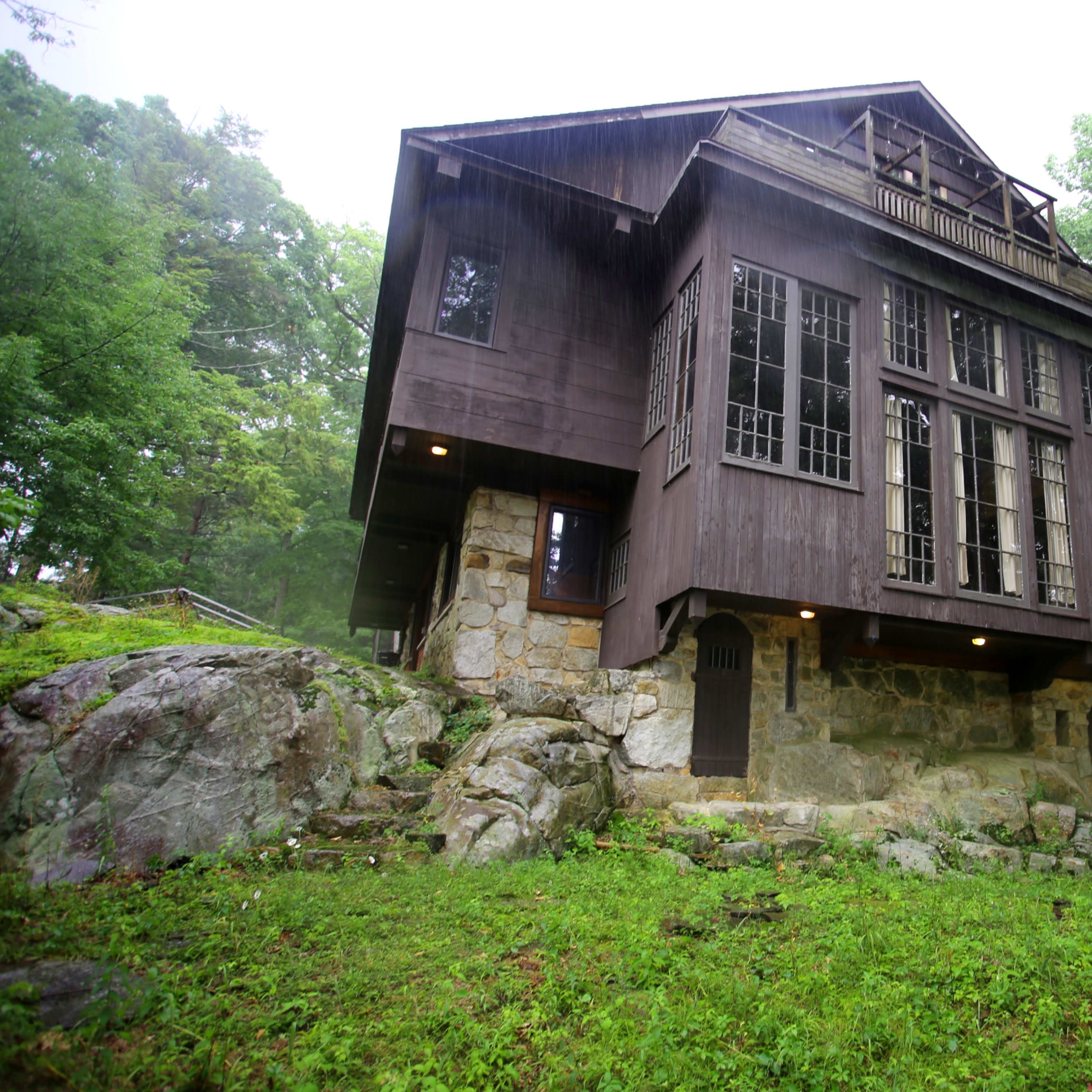 A three-story wooden house sits on a rocky foundation surrounded by dense greenery.