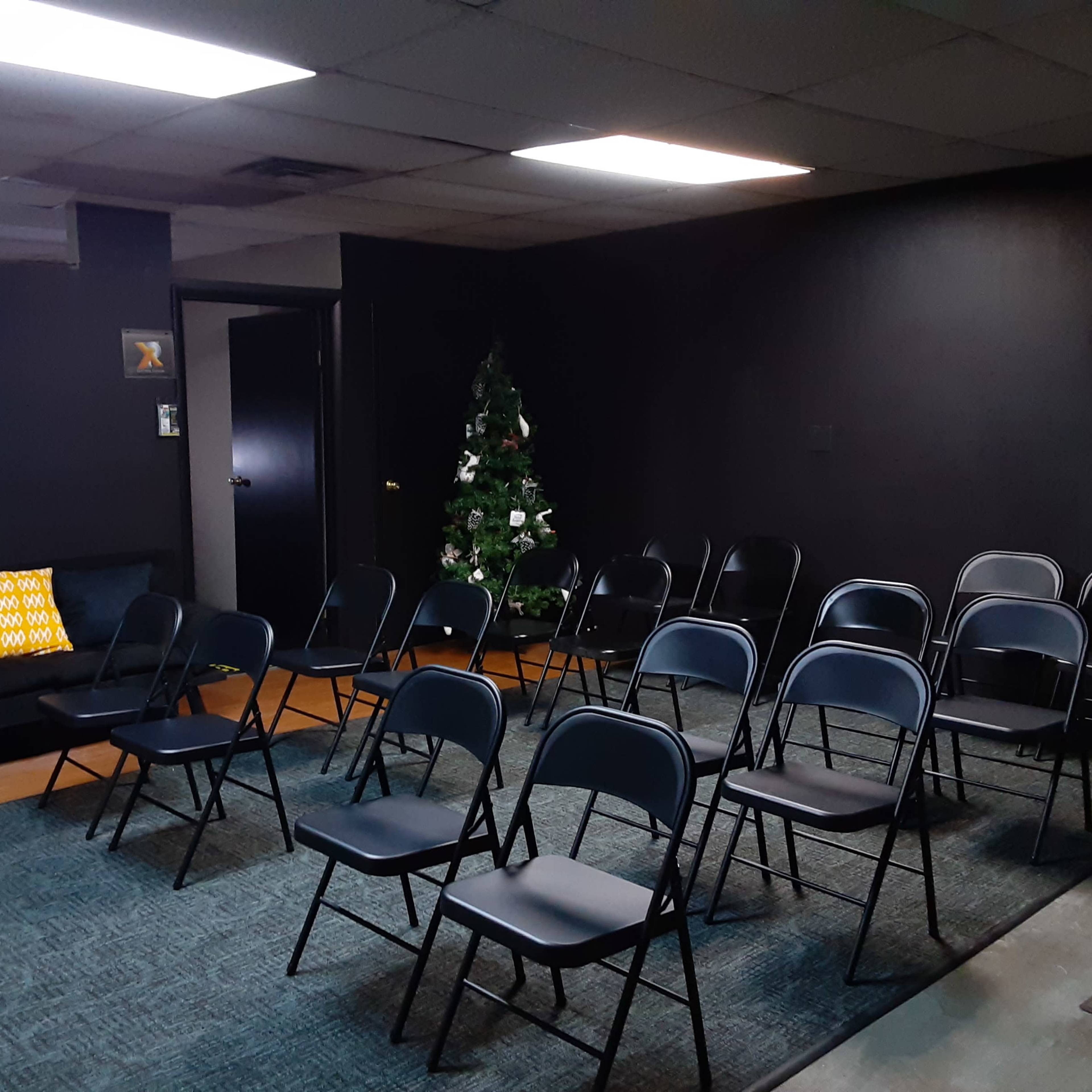 A sparsely furnished room with black walls, a few rows of folding chairs, and a decorated Christmas tree in the corner.