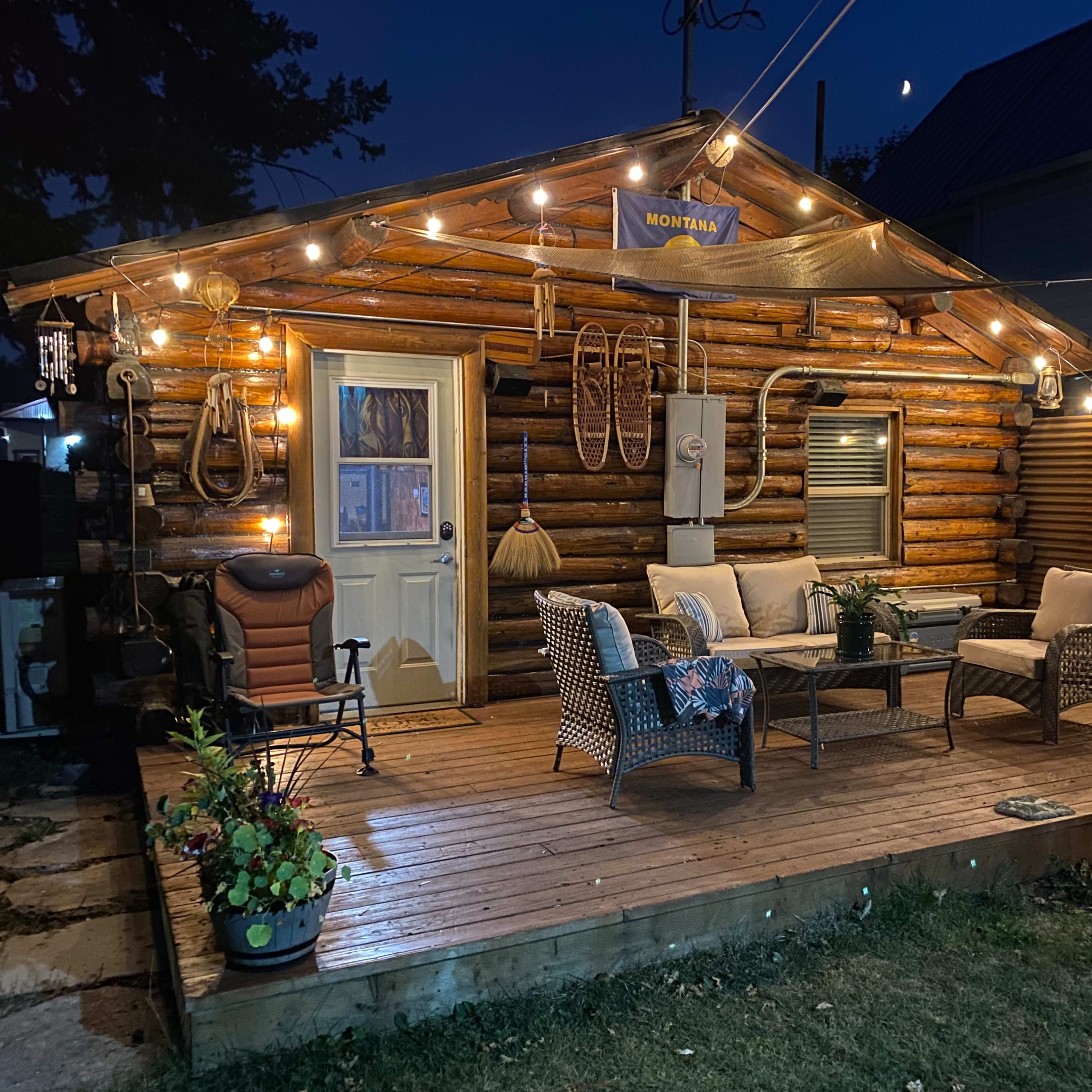 A log cabin with a wooden deck is illuminated by string lights, featuring outdoor furniture and potted plants.