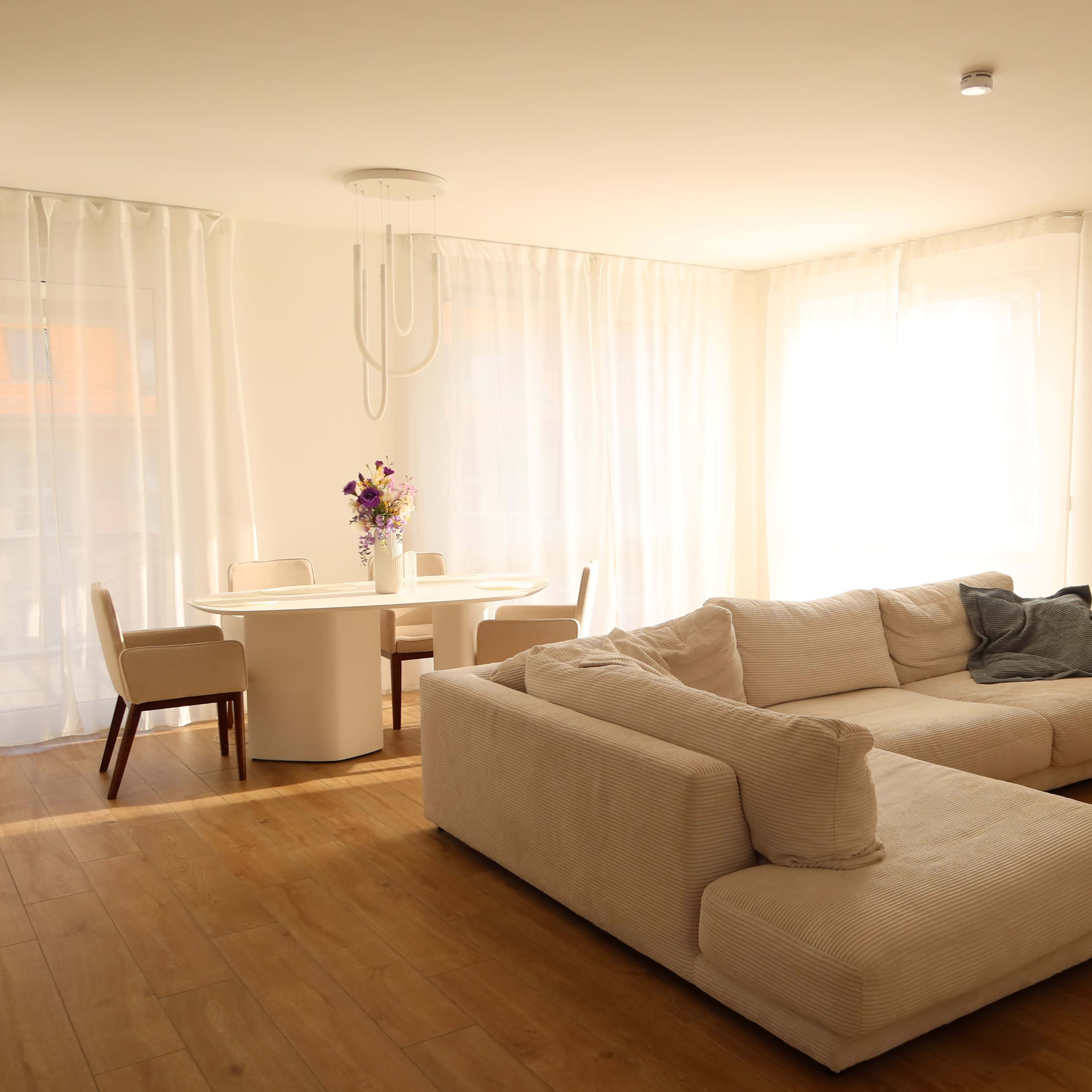 A bright, minimalist living space with a large beige sofa, a round dining table, and white curtains letting in natural light.