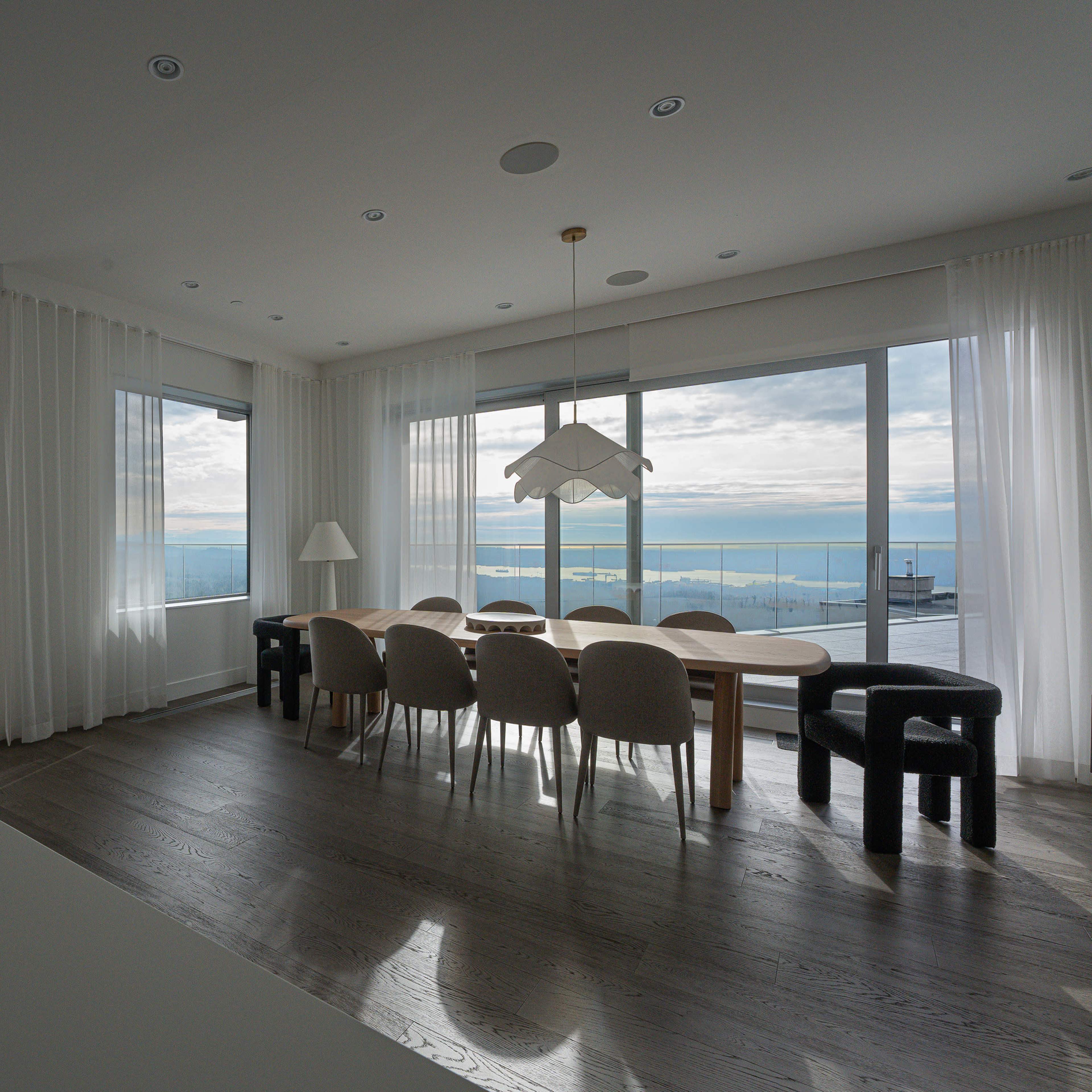 A spacious dining area features a large wooden table surrounded by upholstered chairs, with large windows offering a view of the water and sky.