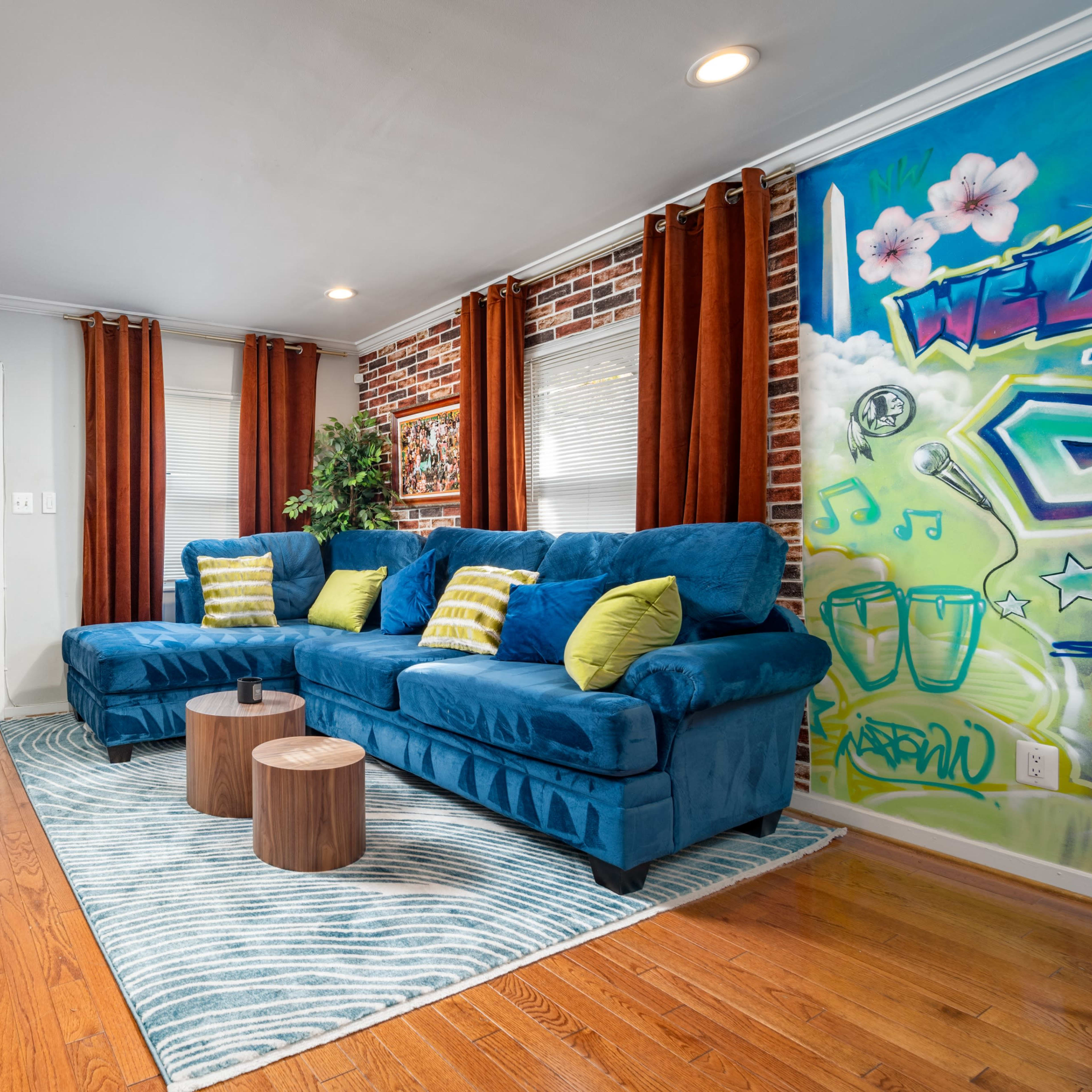 A blue sofa with yellow pillows is positioned in a living room featuring a brick wall, an abstract mural, and wooden furniture.