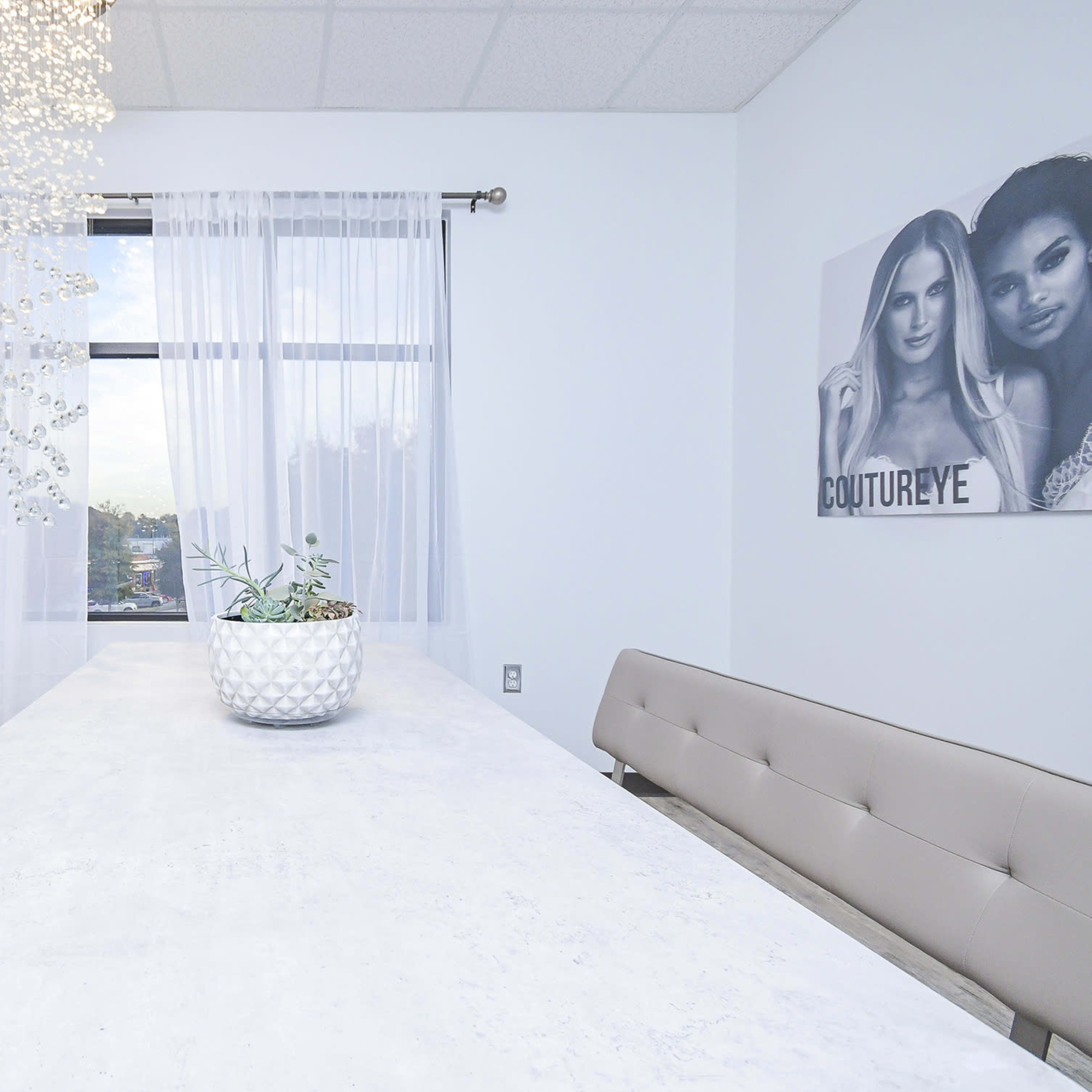 A modern dining or meeting room with a long white table flanked by two light-colored benches, large windows with sheer curtains, and a decorative chandelier.