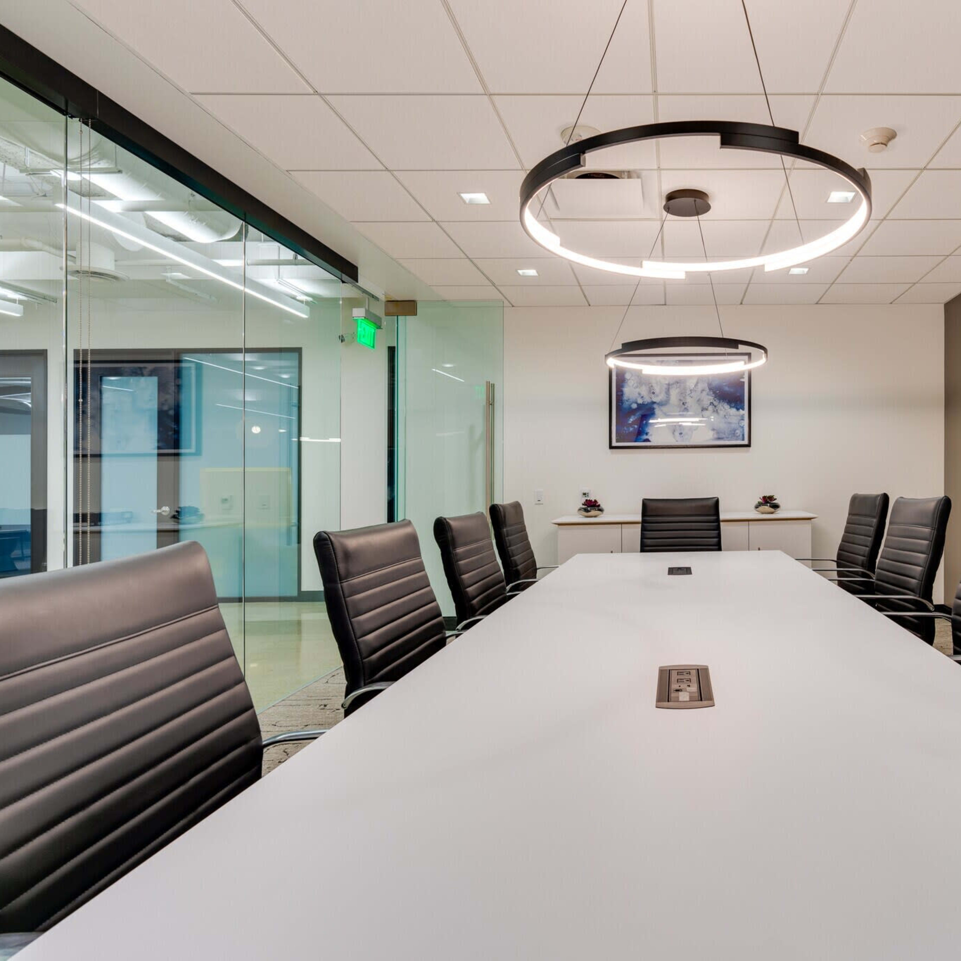 A long conference table with black leather chairs is positioned in a modern meeting room featuring overhead light fixtures and a wall-mounted artwork.