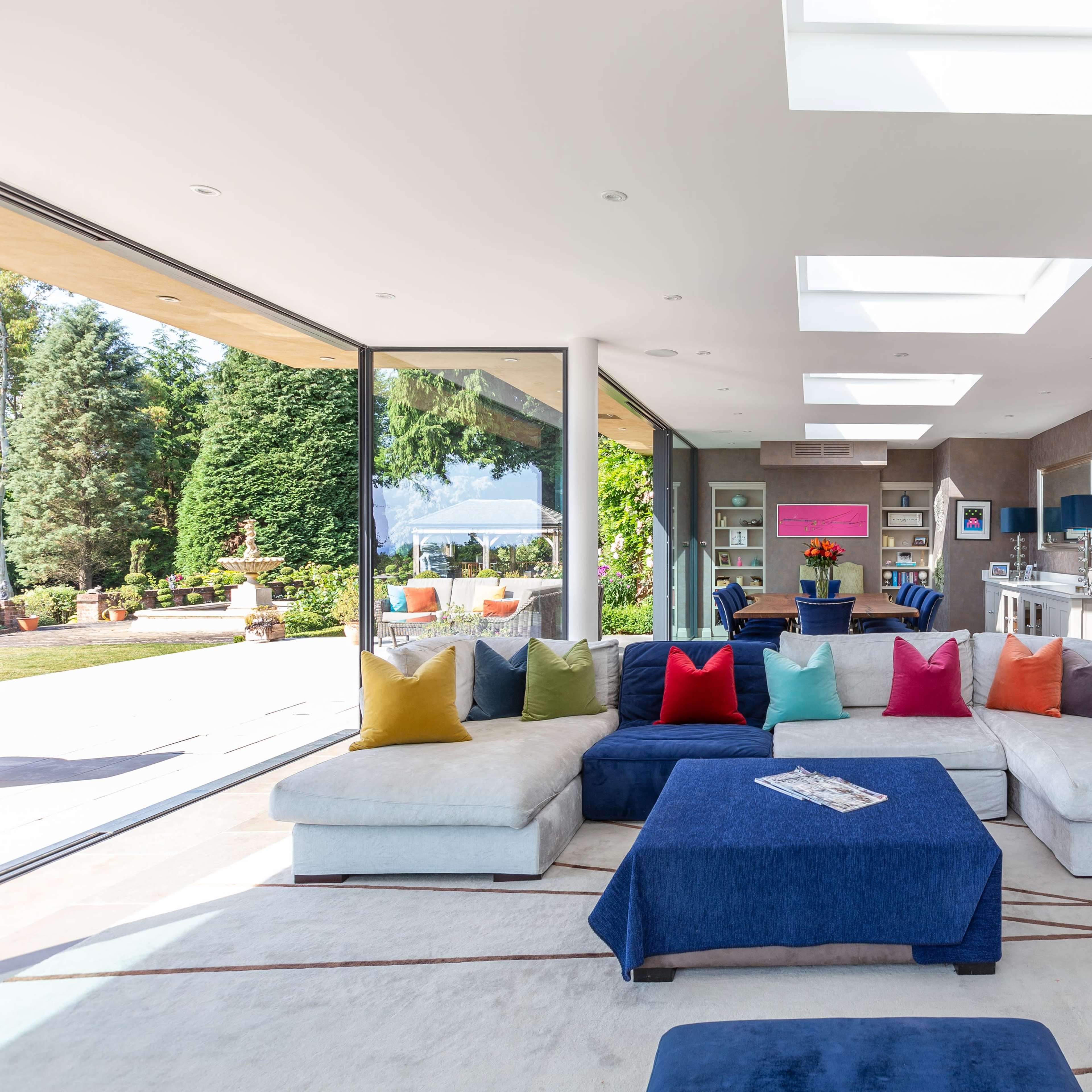 A spacious, modern living room with a large sectional sofa adorned with colorful cushions, overlooking a garden through expansive glass doors.