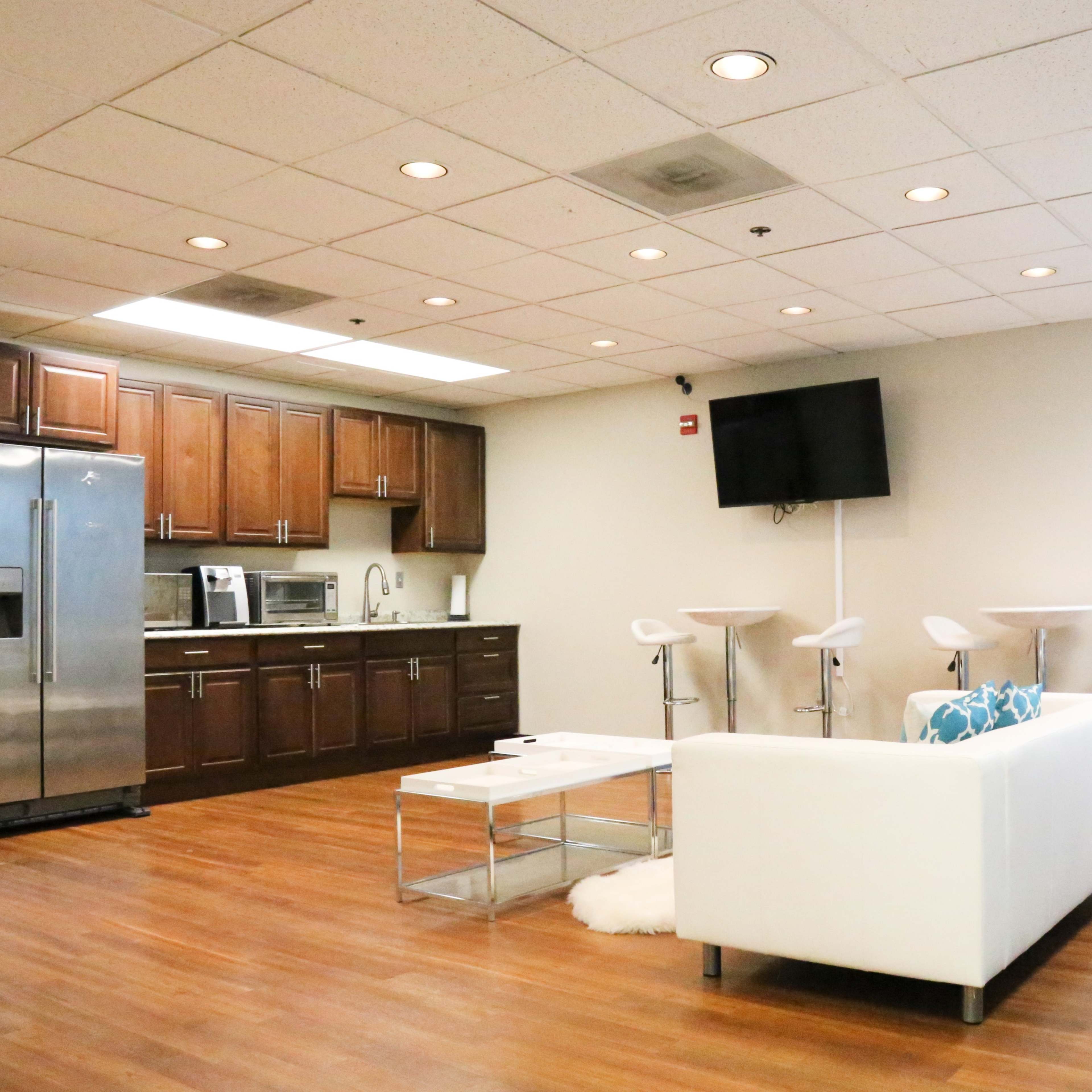A modern break room with a kitchen area, a white sofa, bar stools, and a wall-mounted TV.