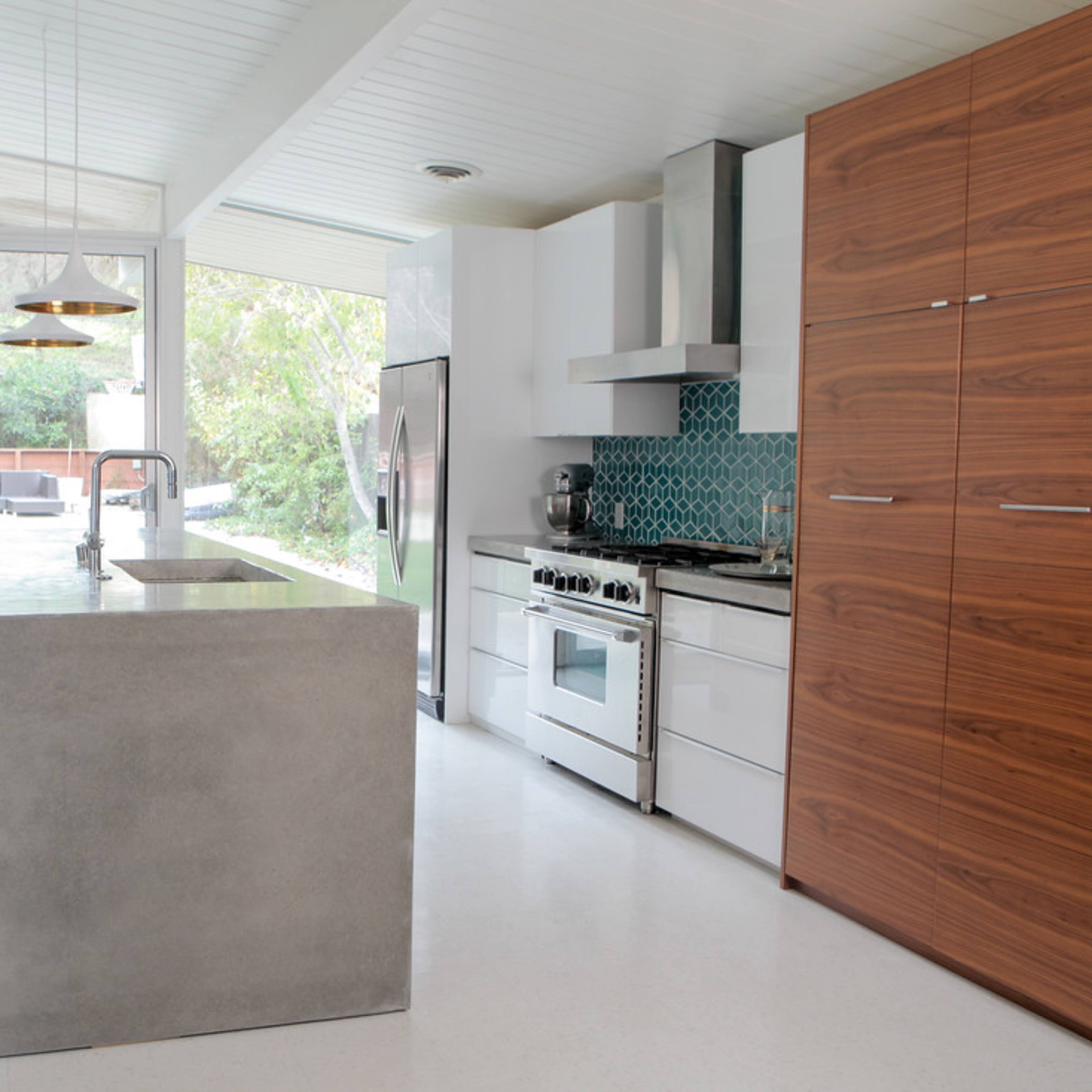 A modern kitchen featuring a concrete island, stainless steel appliances, and wooden cabinetry, with large windows facing an outdoor area.