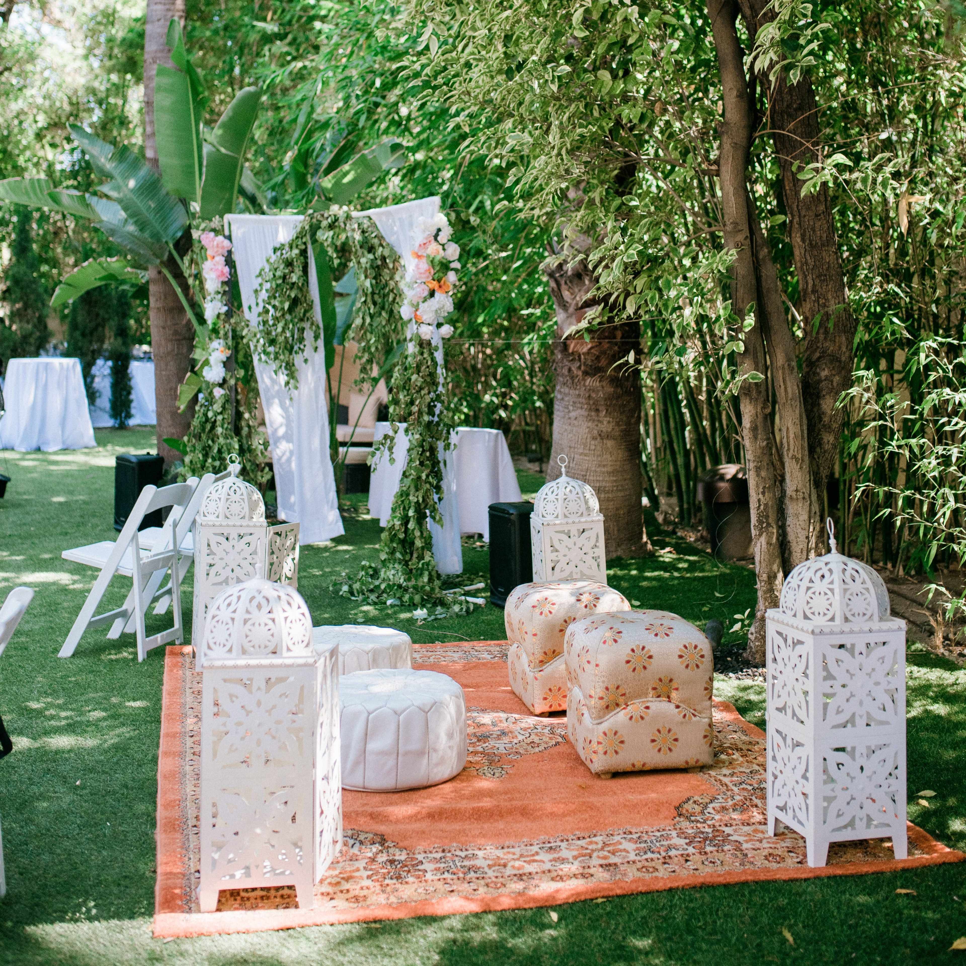 The scene features a beautifully decorated outdoor space with white ornate seating, an orange rug, and a floral backdrop surrounded by greenery.