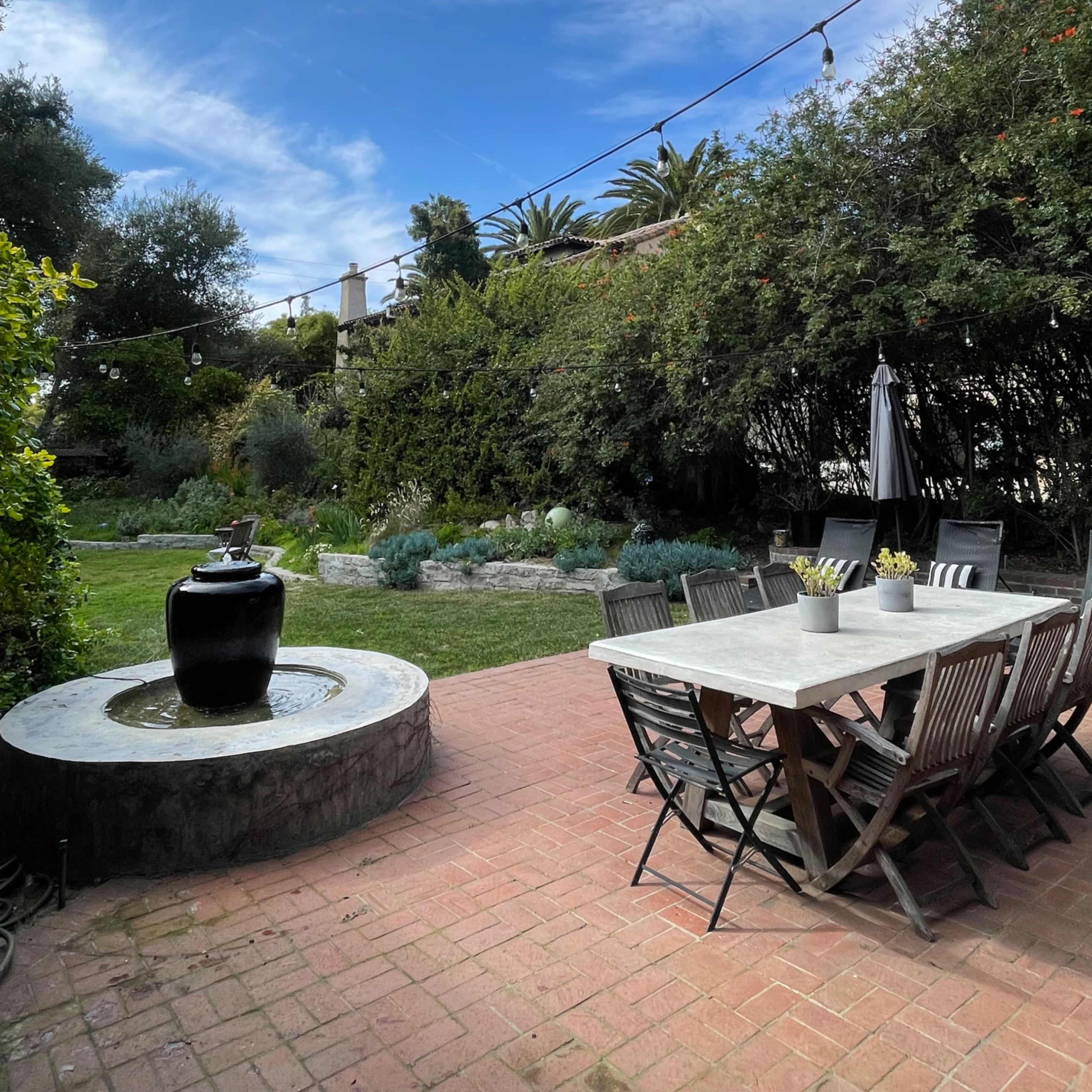 A patio surrounded by greenery features a marble-topped dining table and a black water fountain under a blue sky.