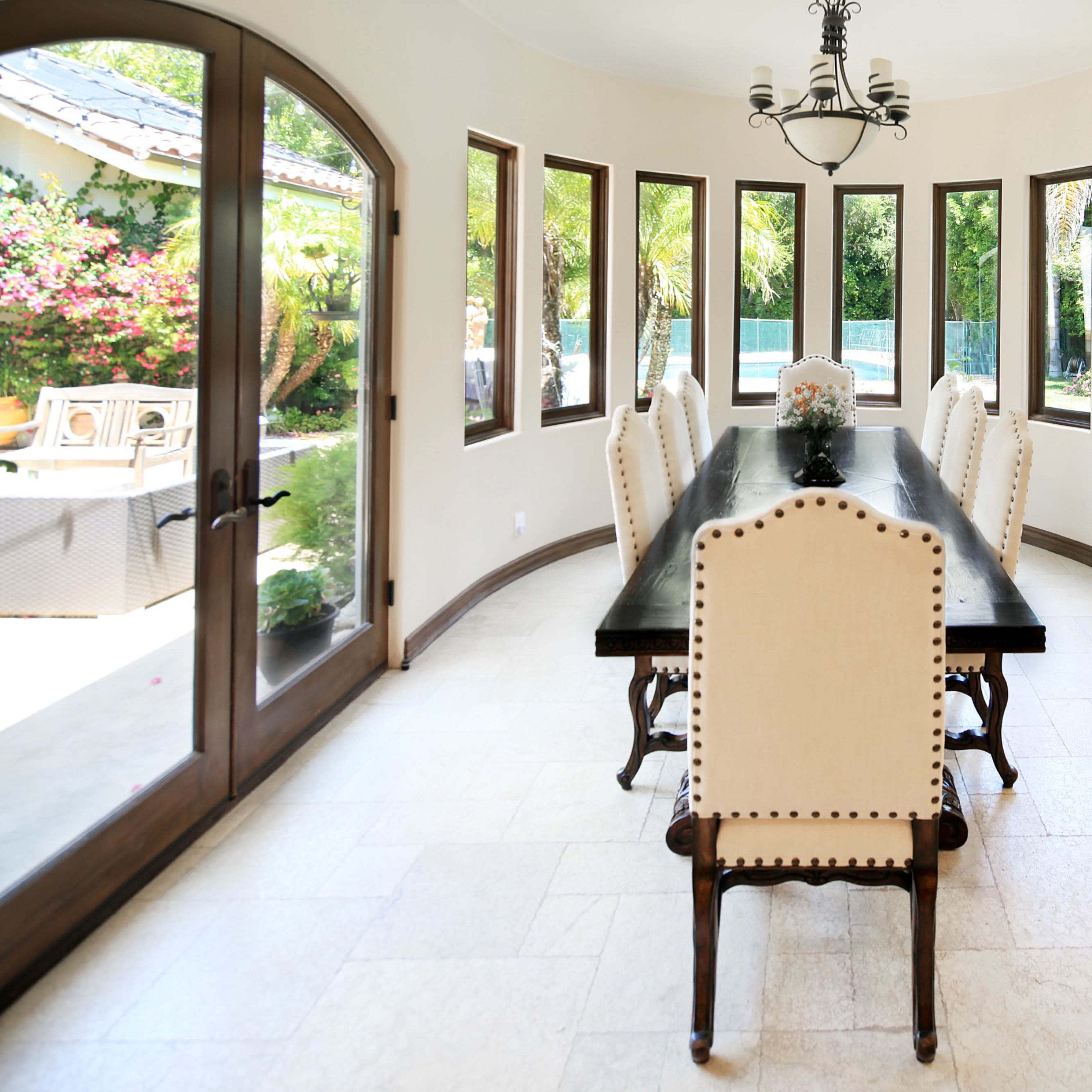 A spacious dining area features a long black table surrounded by twelve light-colored chairs and large windows offering views of a lush outdoor landscape.