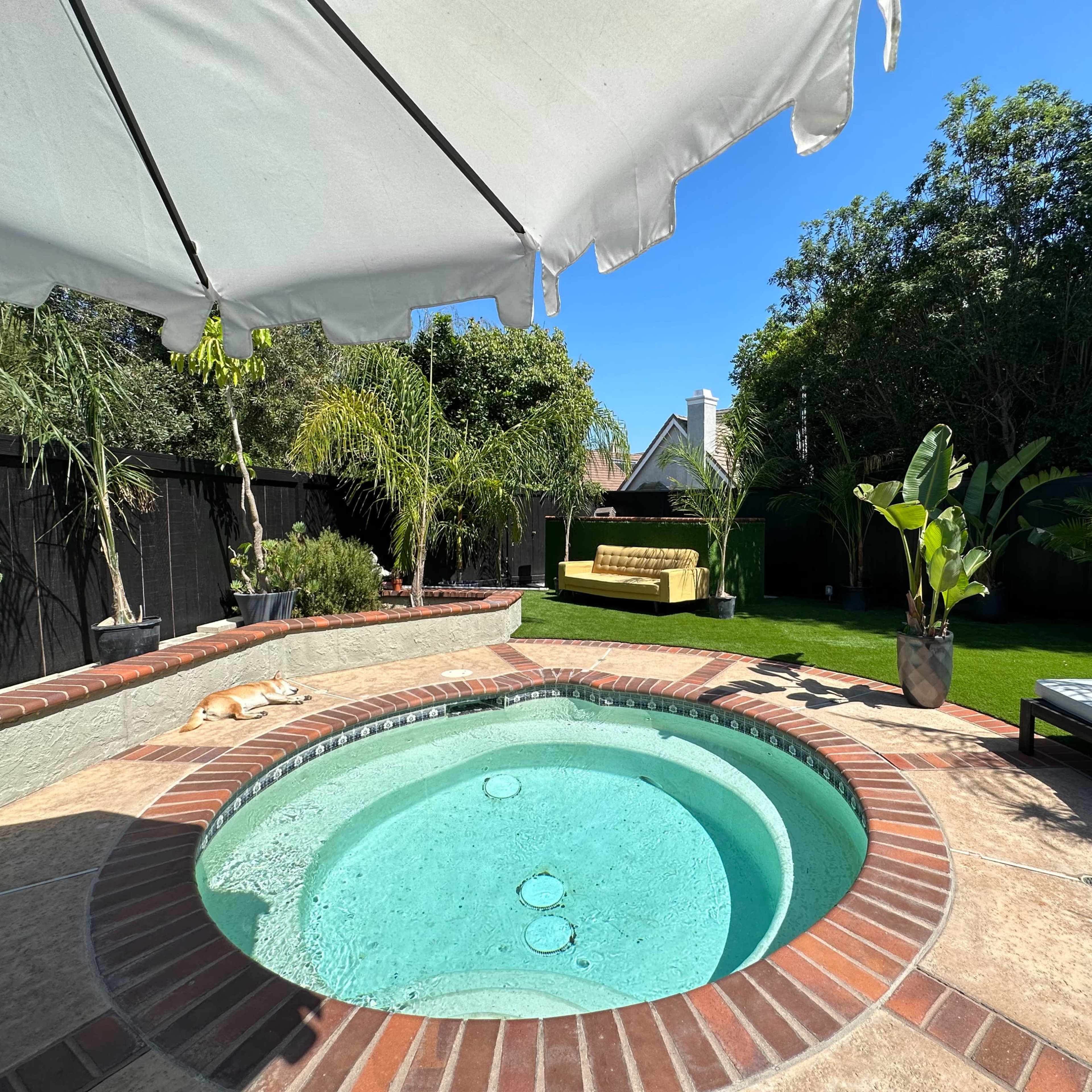 A circular spa is set within a landscaped backyard featuring a grassy area, potted plants, and a covered seating space.