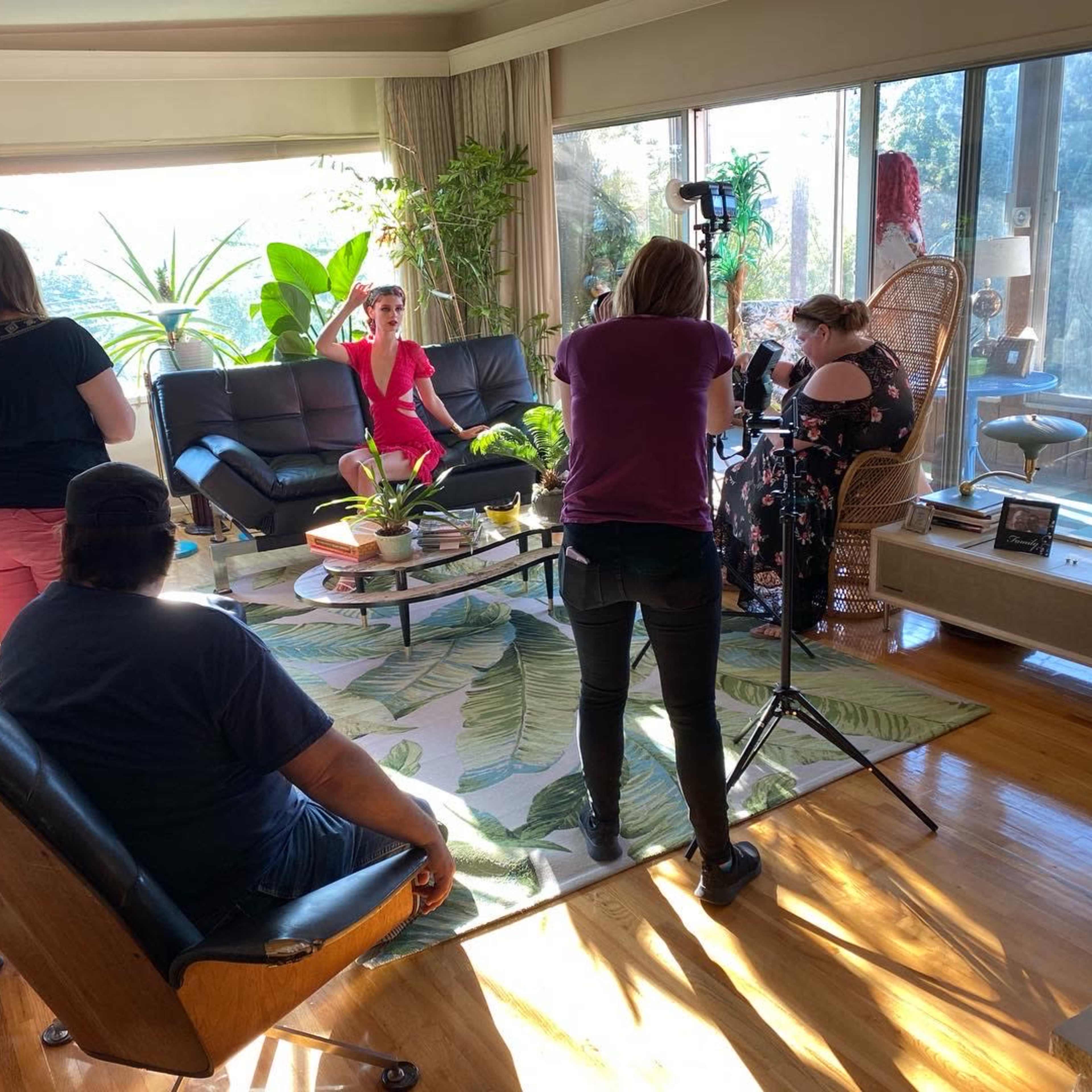 A group of people is gathered in a brightly lit living room, with one person in a red dress posing for a photo while others work with cameras and equipment.