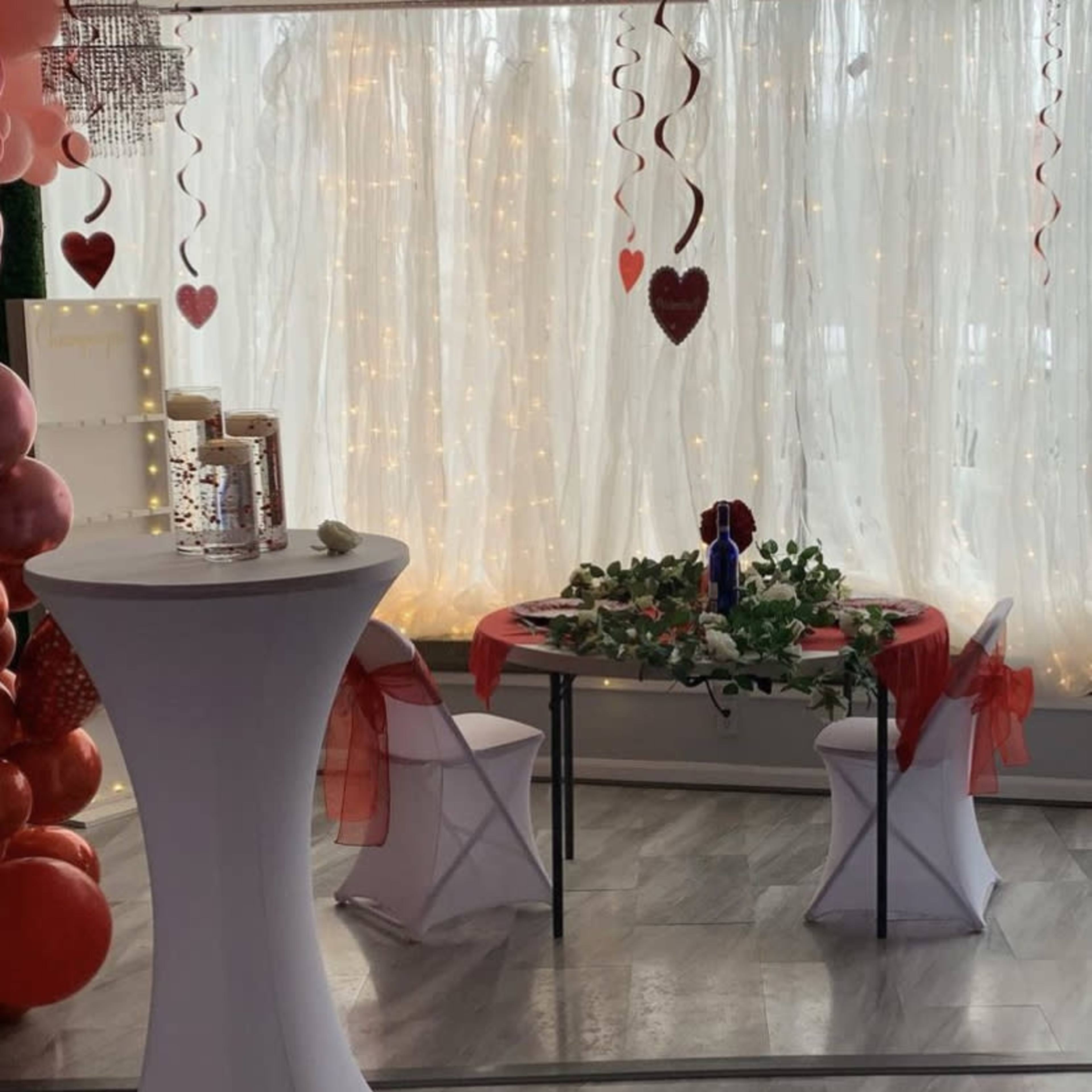 A decorated table with a floral centerpiece, surrounded by chairs, is set up in a room adorned with hearts and fairy lights, while candles in glass containers are placed on the floor nearby.