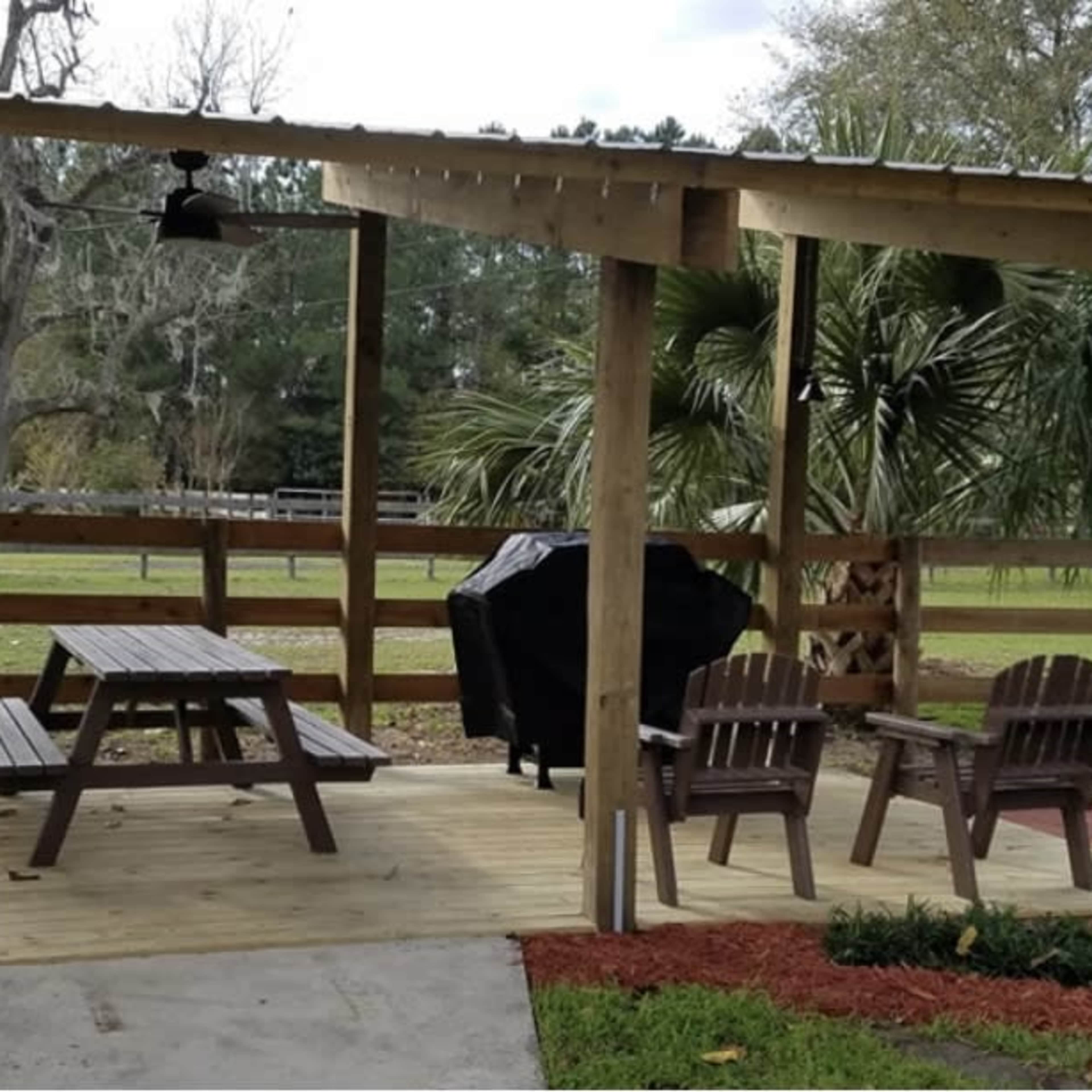 A wooden patio structure with a picnic table and two chairs, alongside a barbecue grill and a landscaped area with grass and mulch.