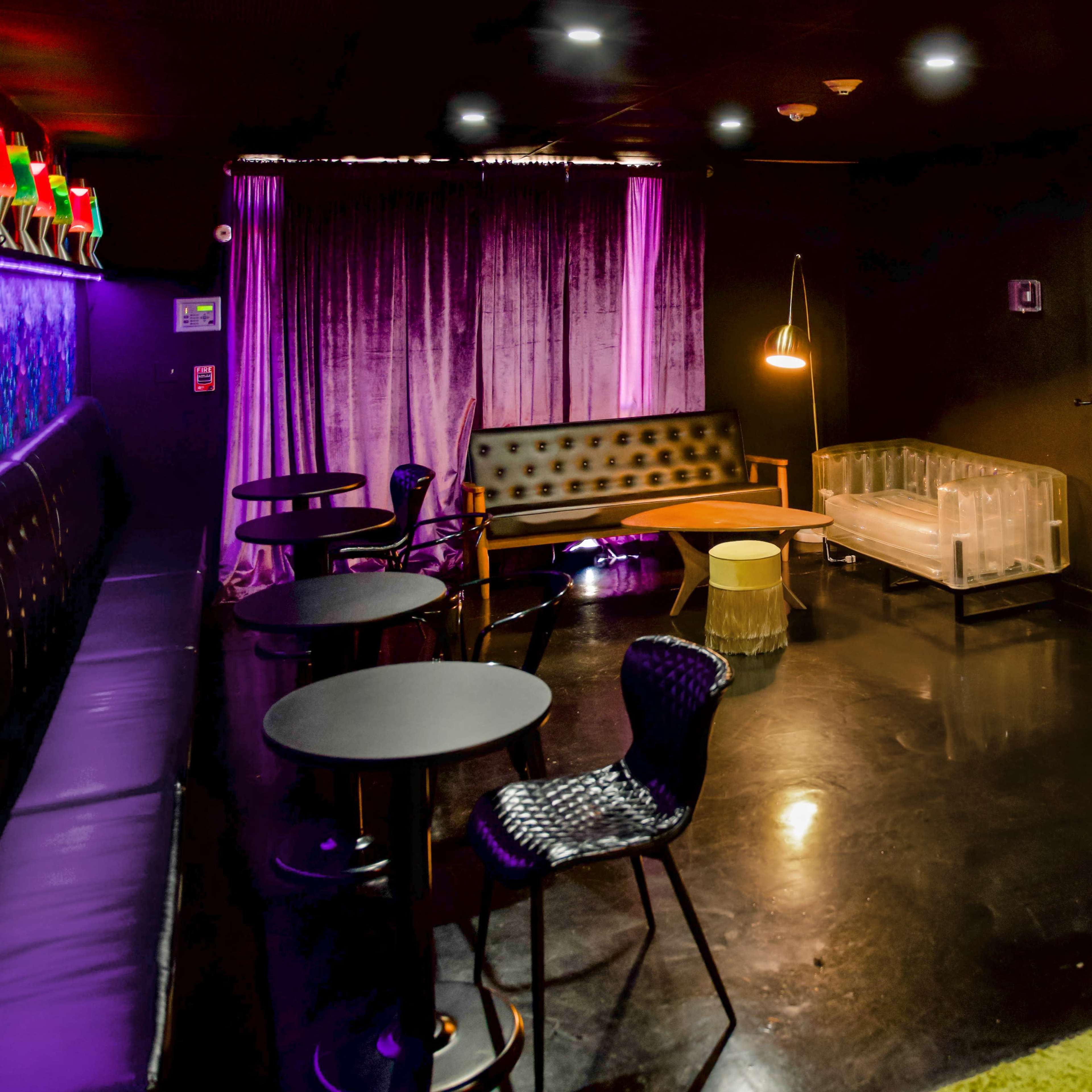 A stylish lounge area with black tables, mixed seating options, and purple curtains, illuminated by soft lighting.