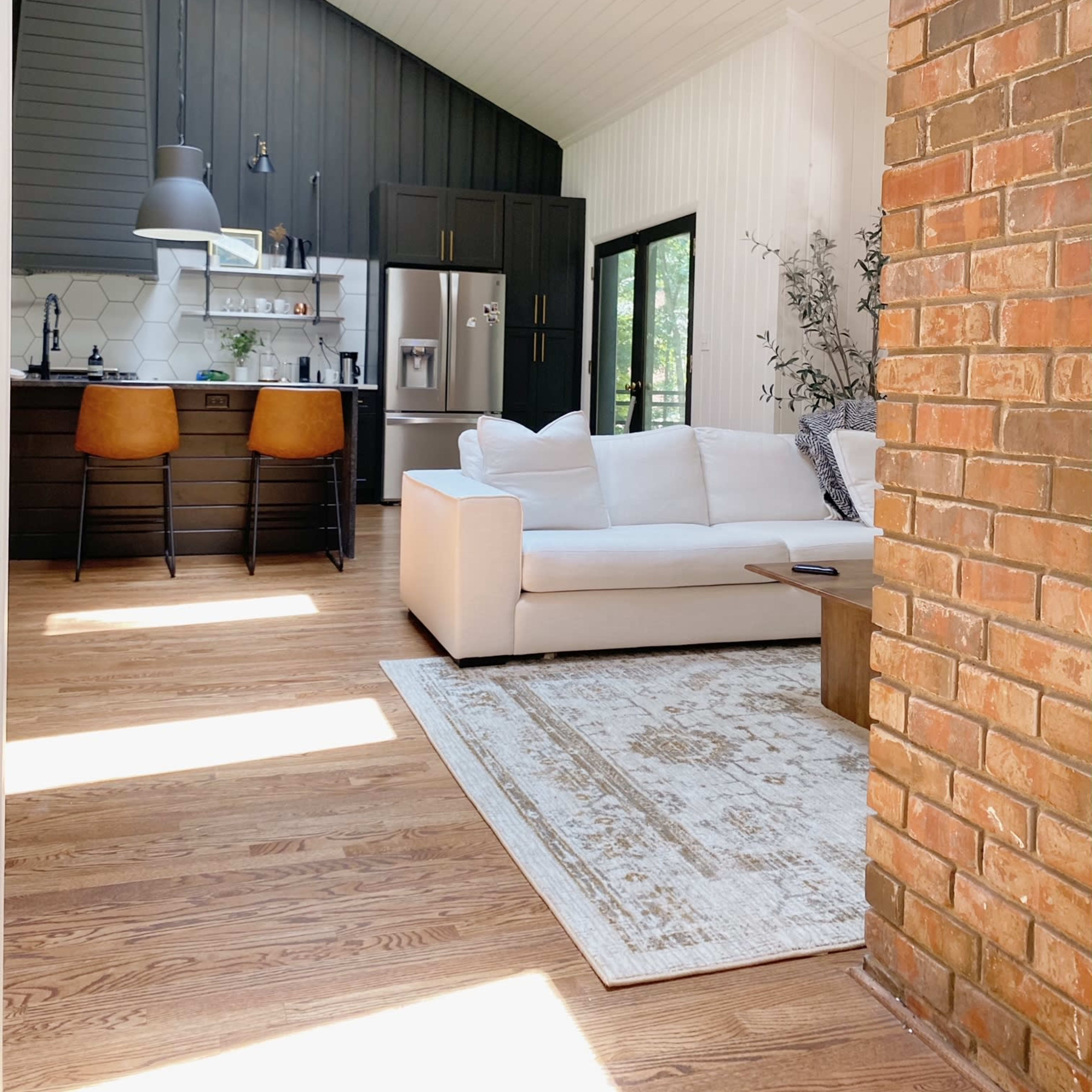 A modern living space with a light-colored sofa, a wooden table, and a kitchen area featuring dark cabinets and large windows for natural light.