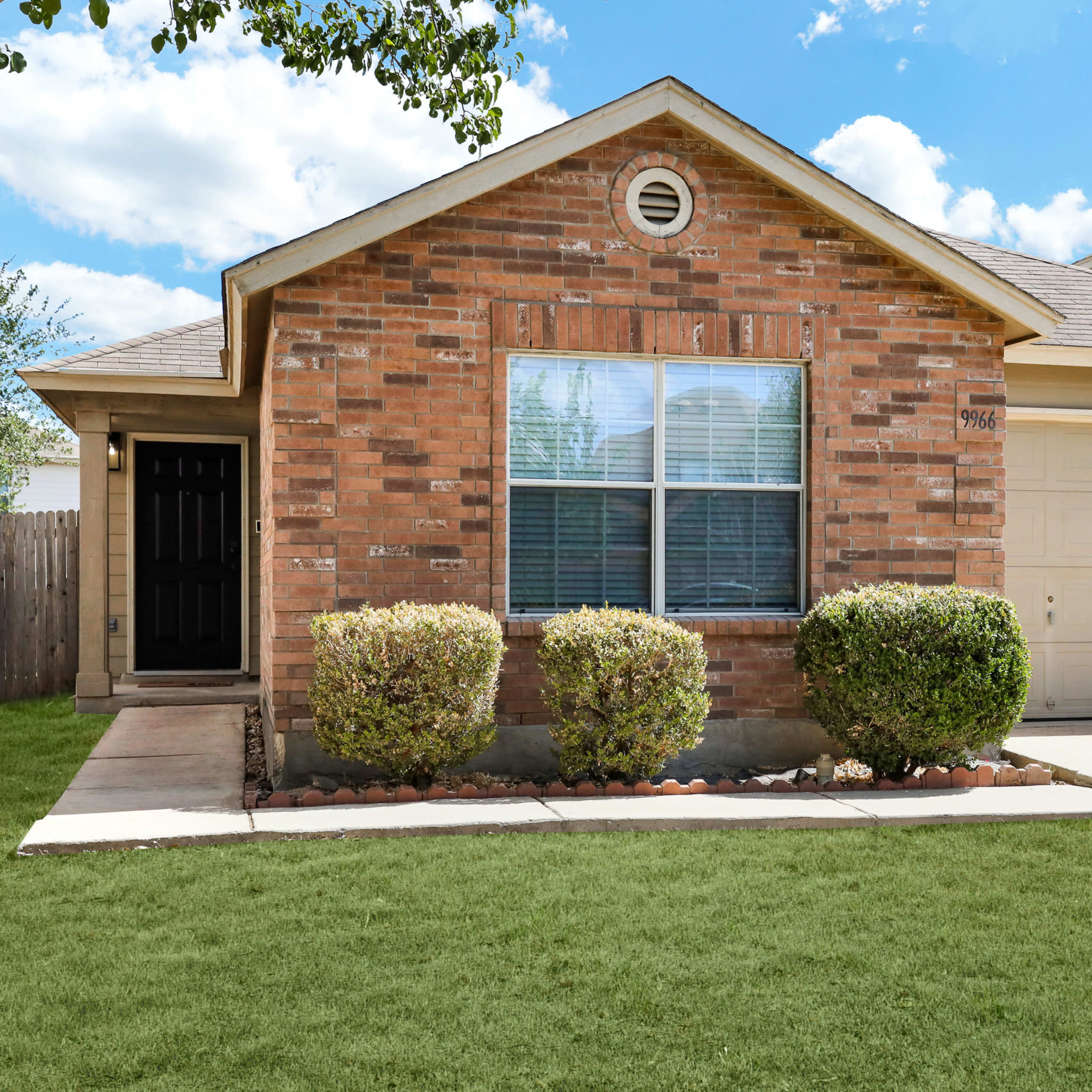 A single-story brick house with a front lawn, two shrubs by the entrance, and a single-car garage.