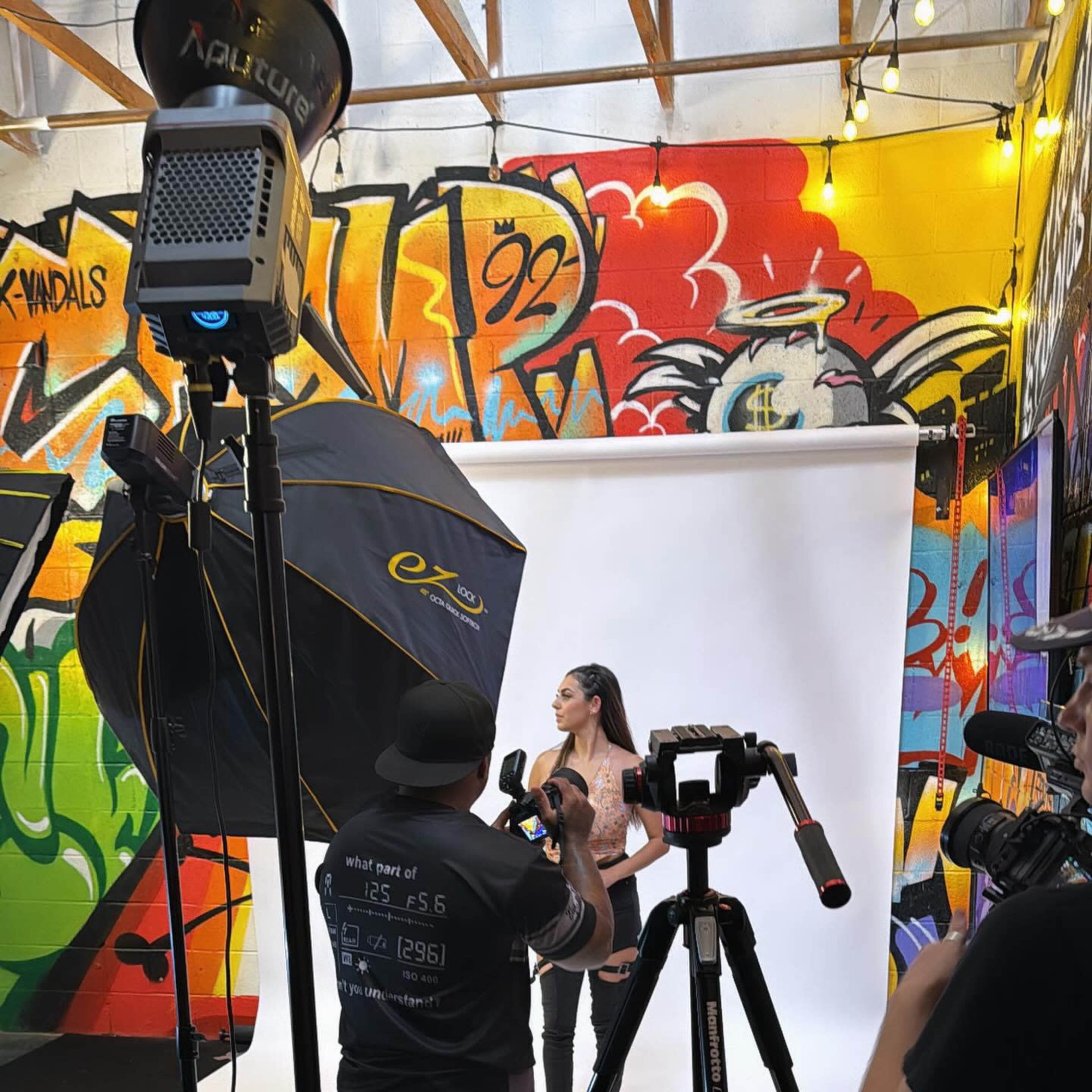 A model poses in front of a white backdrop while photographers and lighting equipment surround her in a graffiti-decorated studio.