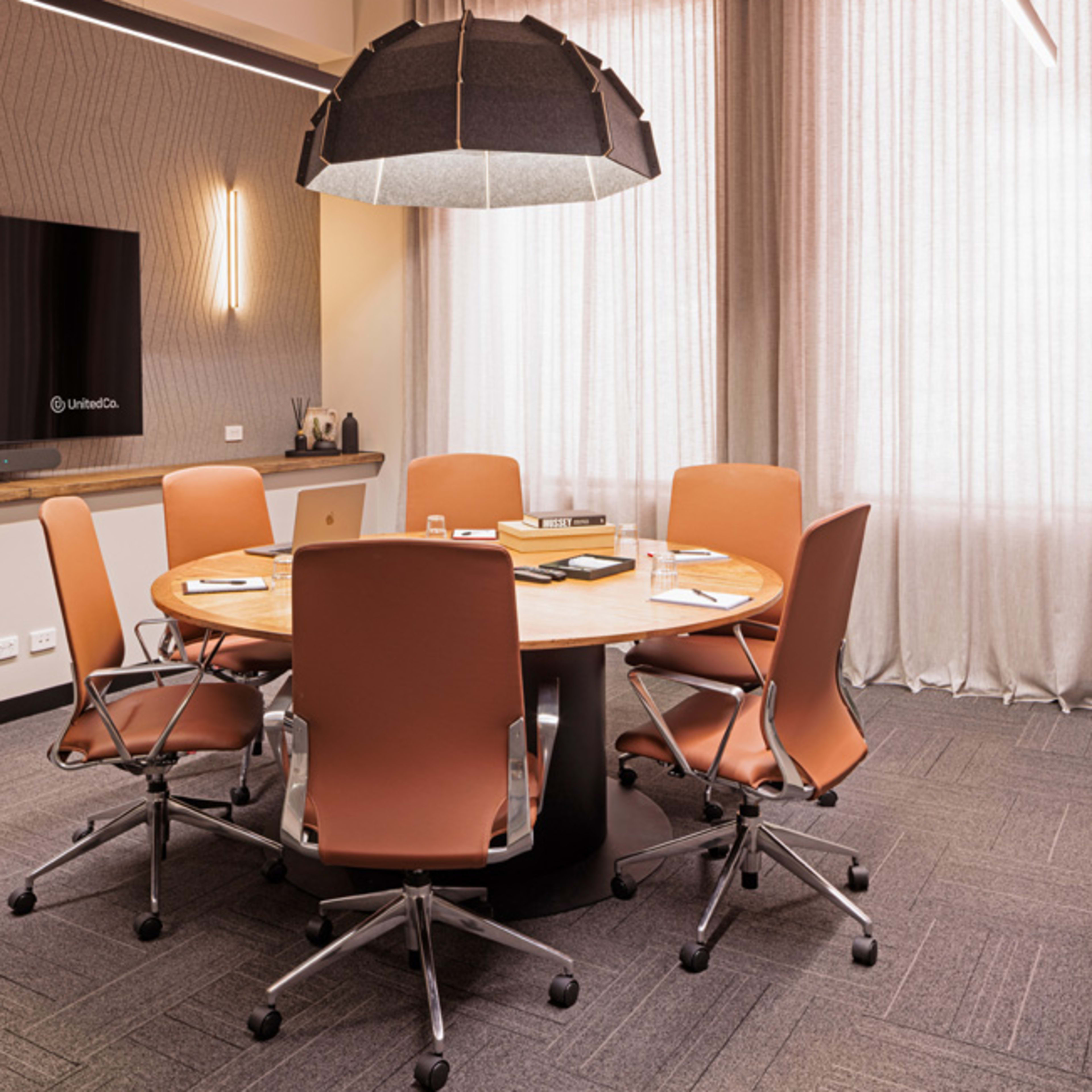 A modern conference room features a round wooden table surrounded by six ergonomic chairs, with a large screen on the wall and natural light filtering through sheer curtains.