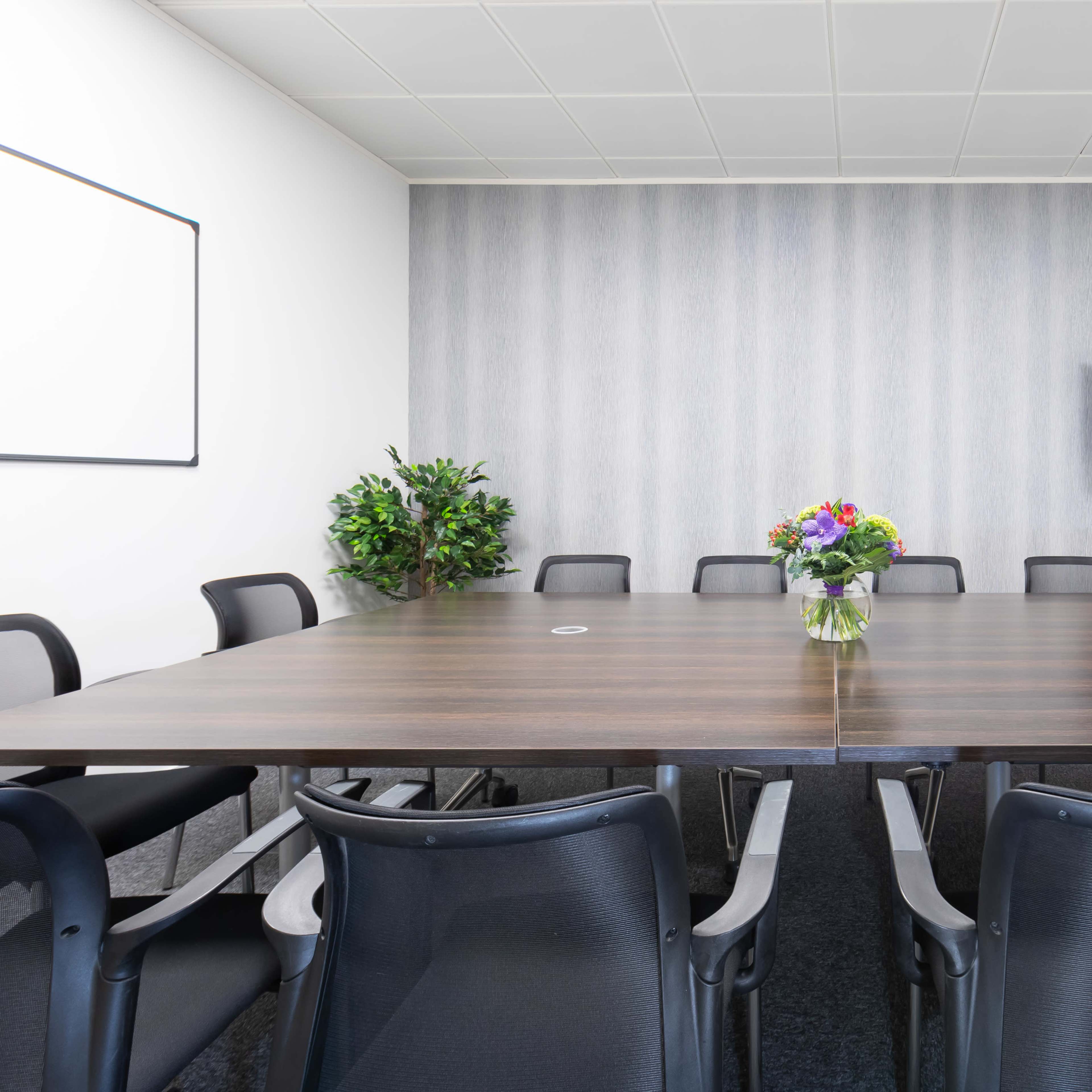 A conference room is set up with a large wooden table surrounded by black chairs, featuring a TV on a stand and a vase of flowers in the center.