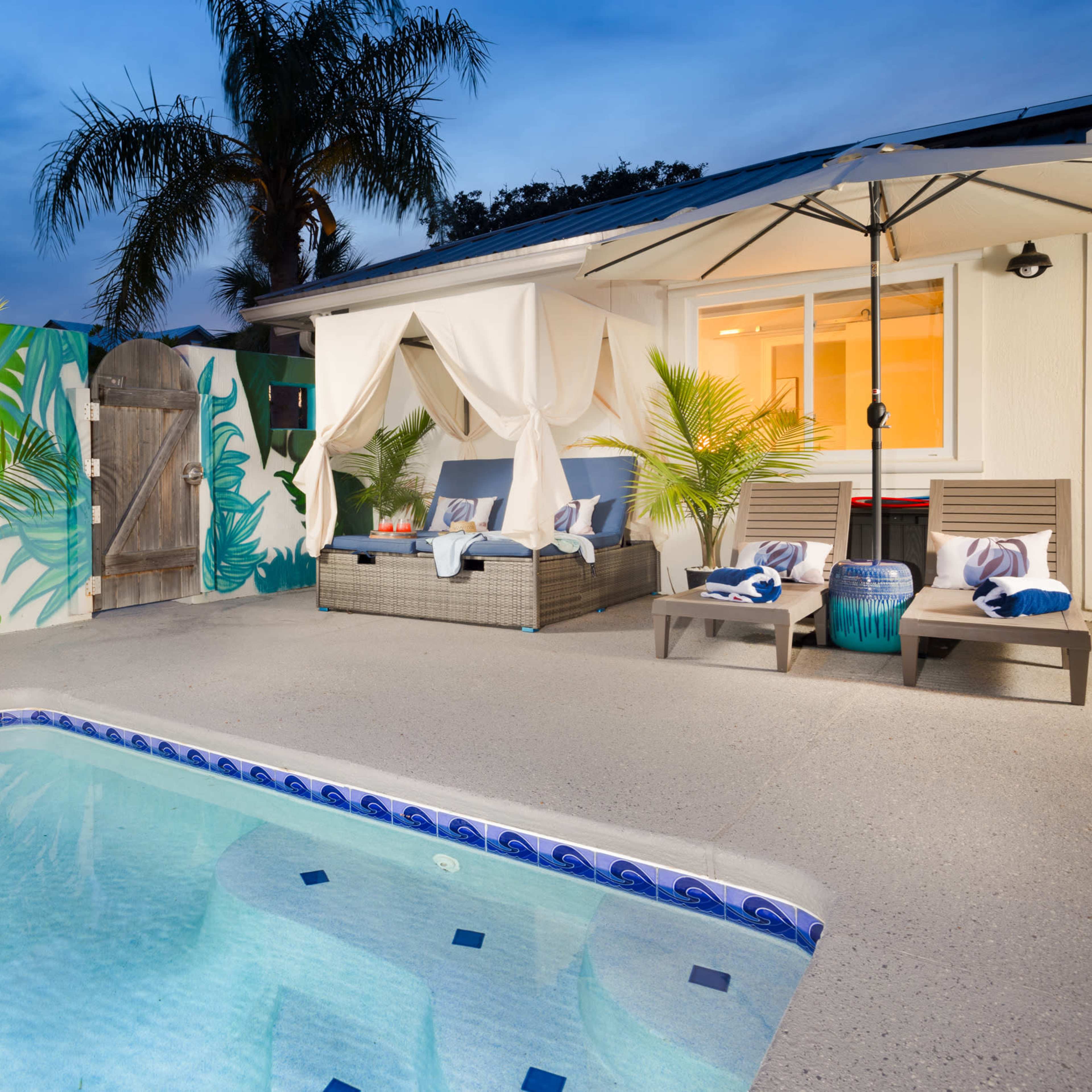 A pool area with a mural of a bird on the wall, lounge chairs with towels, and a shaded seating area beside the water.