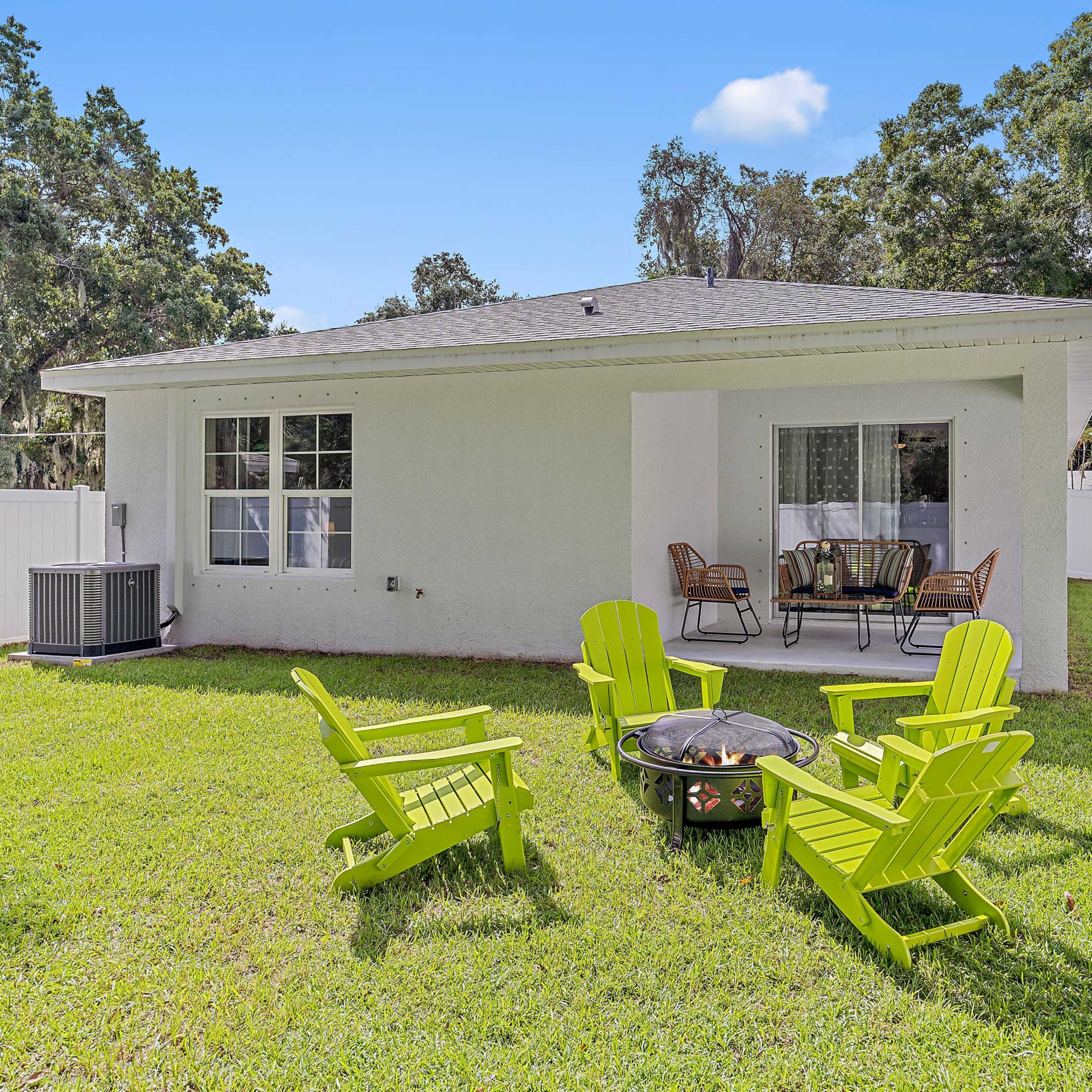 A back yard with a fire pit surrounded by four green chairs, a small table, and a view of a house with large windows.